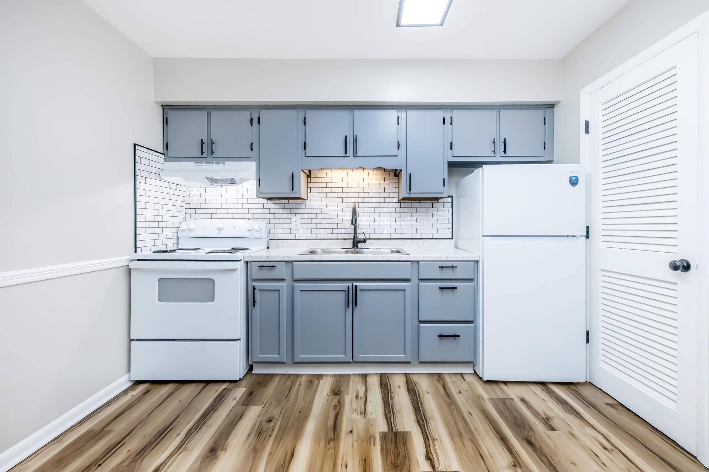 A modern kitchen featuring gray cabinets, a white stove, a sink with a black faucet, and a white refrigerator. The backsplash consists of white tile, and the floor has a wood-like finish. Bright overhead lighting enhances the clean and minimalistic design of the space.