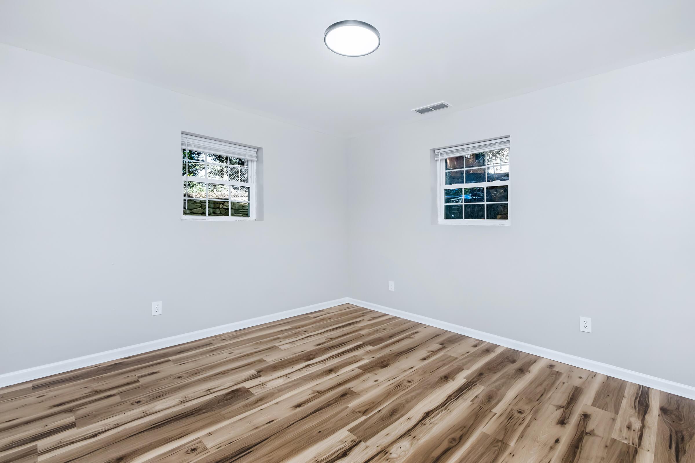 A bright, empty room featuring light-colored walls, two windows with white blinds, and a ceiling light fixture. The flooring is wooden, showcasing a natural finish. The overall ambiance is clean and modern, with no furniture or decorations present.