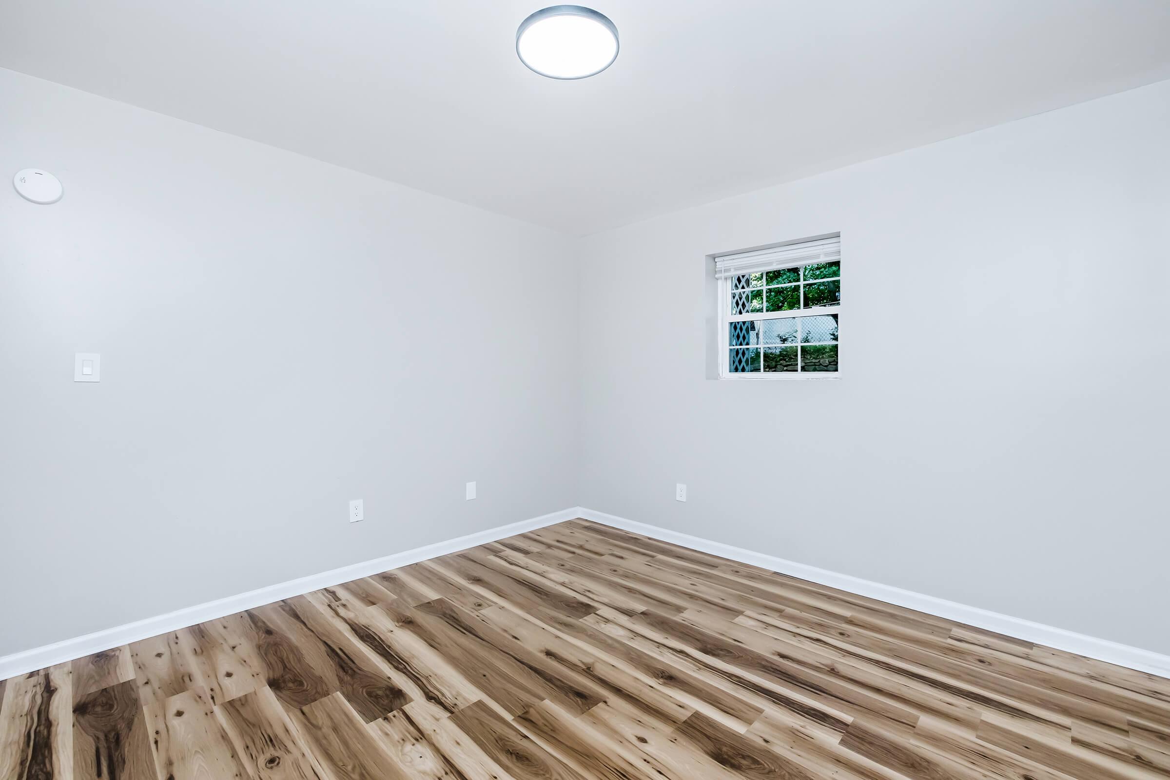 A spacious room with light gray walls and a smooth wooden floor. There is a window with a white frame allowing natural light, and a modern ceiling light fixture. The room is empty, creating a clean and minimalist atmosphere.