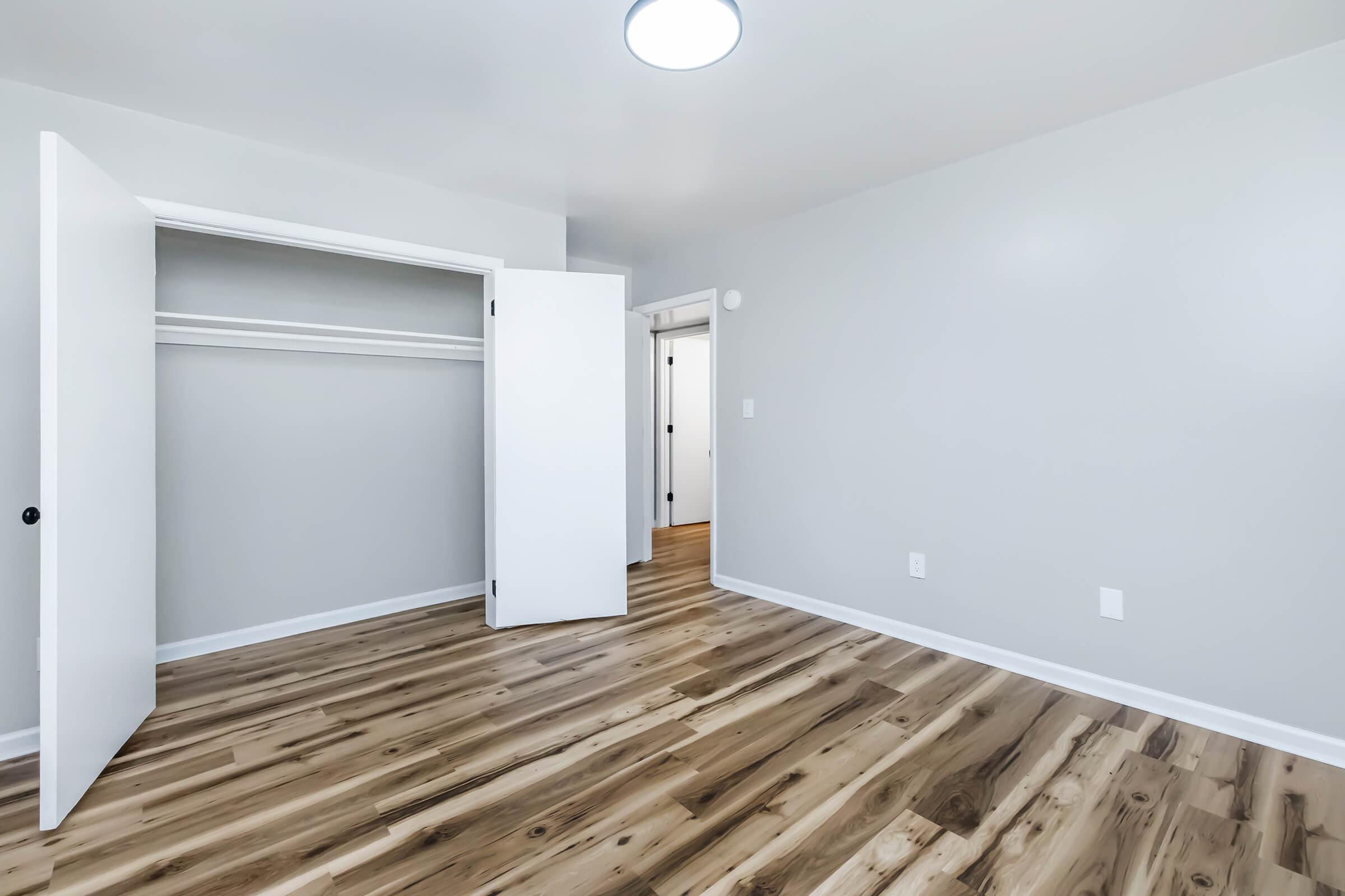 A clean, empty bedroom featuring light-colored walls, a wooden floor, and a large closet with double doors. The room is well-lit by a ceiling light and has a doorway leading to another space, suggesting a modern, open layout.