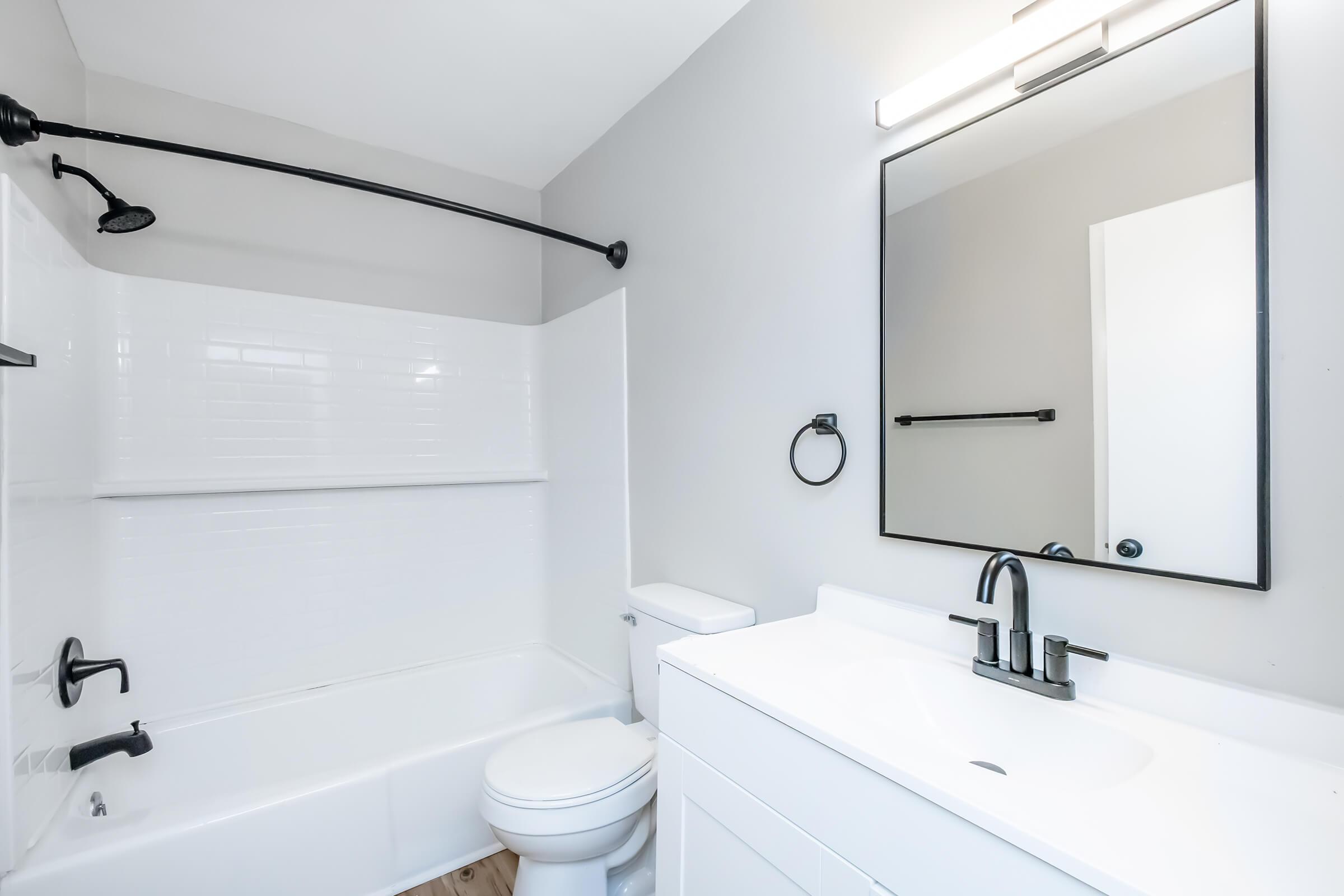A modern bathroom featuring a white bathtub with a shower, a sleek black showerhead, a white vanity with a sink, a large mirror above the sink, and a toilet. The walls are painted gray, and there is a towel ring on the wall. Natural light is coming from the doorway.