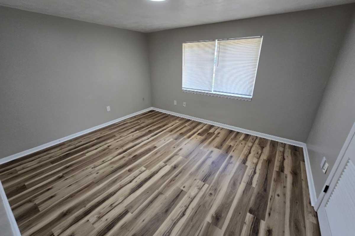 Empty room with light gray walls and a window covered by horizontal blinds. The flooring is made of light wood planks, giving a modern look. The room is well-lit, with natural light coming from the window, and features a simple design with no furniture.