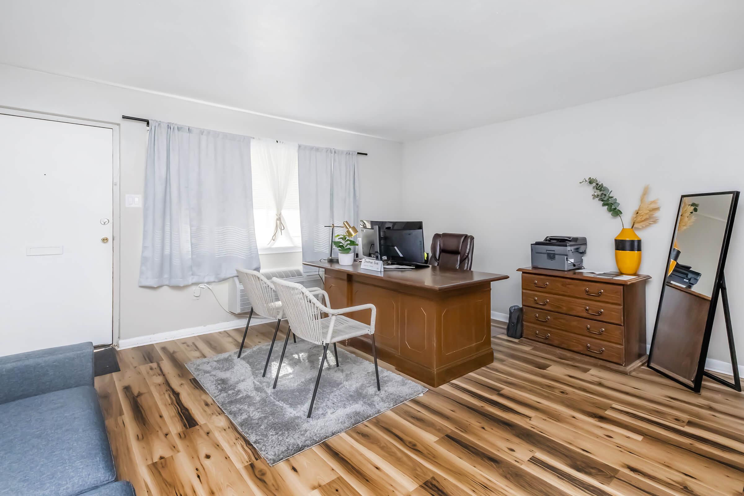A bright and airy home office space featuring a wooden desk with a computer, two white chairs, and a large mirror. Natural light filters through sheer curtains by the window. The floor is covered with light wood planks, and there's a gray area rug under the desk. A plant and printer are visible, adding to the functional setup.