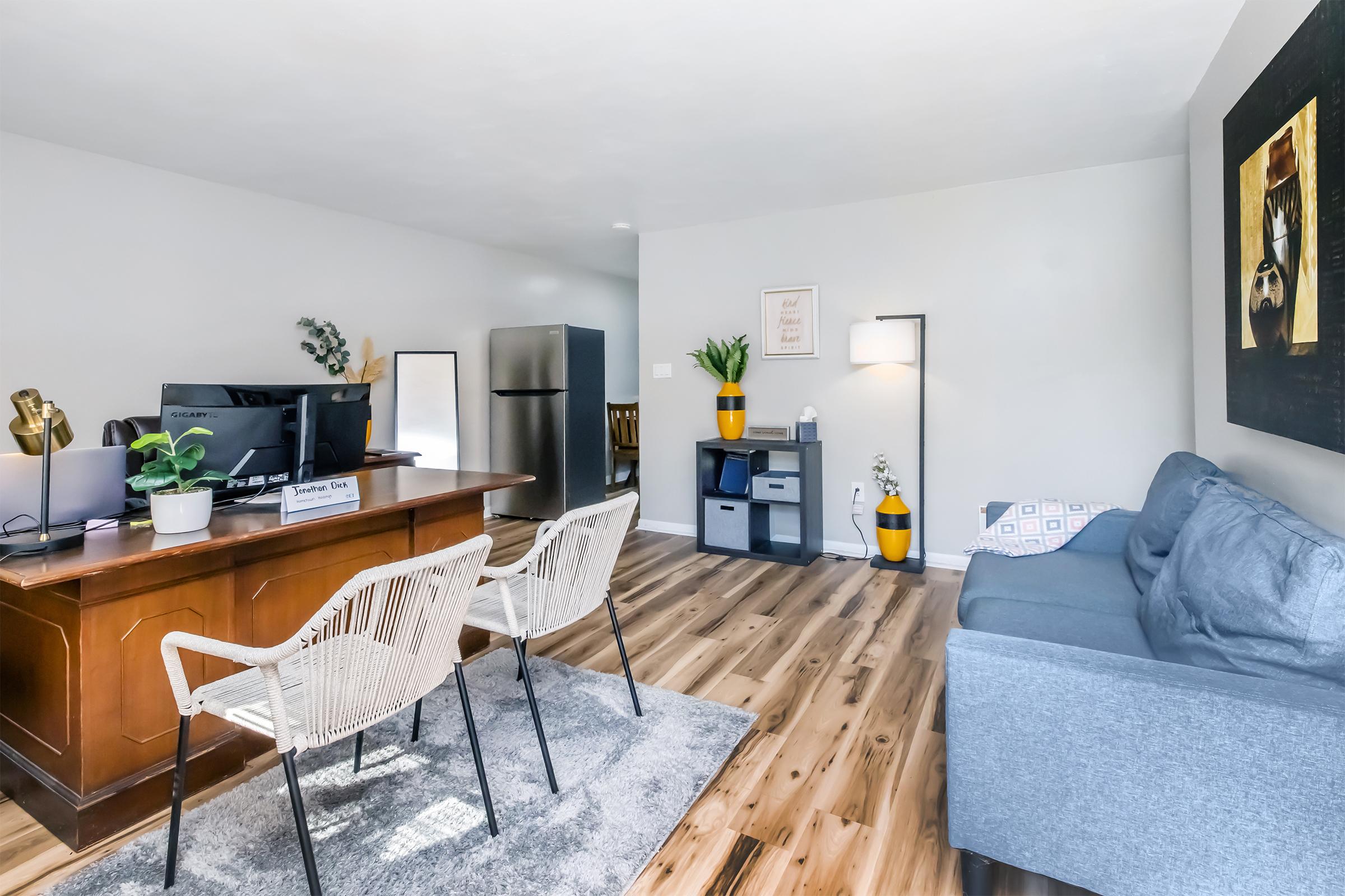 A modern office space featuring a wooden desk with a computer setup, two stylish chairs, a gray sofa, and a small shelving unit. Decorative elements include a potted plant, a floor lamp, and artwork on the wall. The flooring is wooden and the walls are painted light gray, creating a bright and inviting atmosphere.