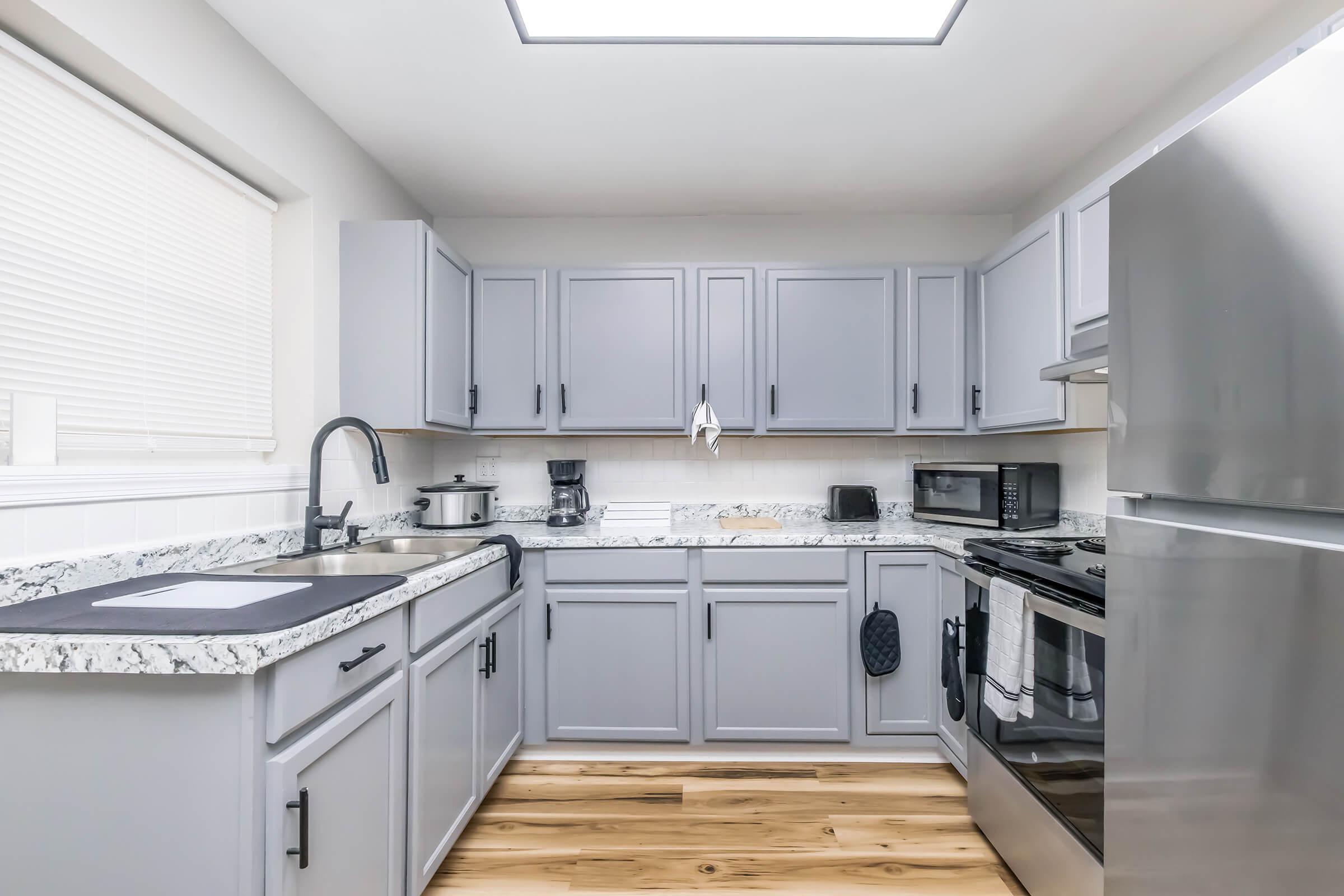 Modern kitchen with gray cabinets, white marble countertops, and stainless steel appliances. A large window provides natural light, while wooden flooring adds warmth. The setup includes a sink, stovetop, microwave, and coffee maker, creating a stylish and functional space for cooking and dining.