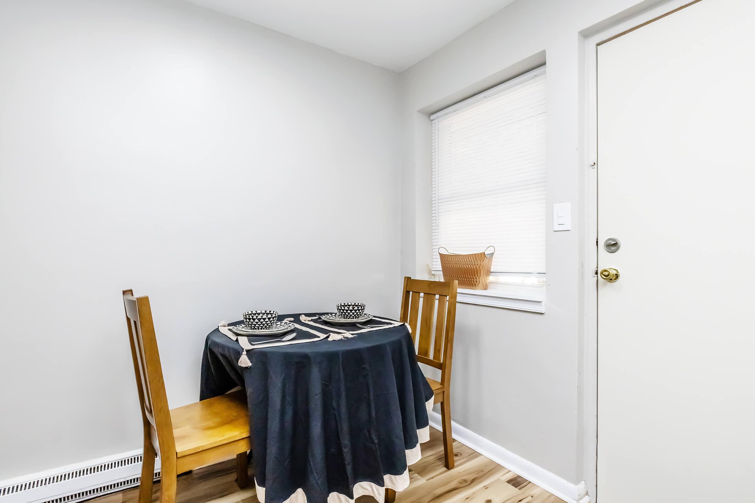 A small dining area featuring a wooden table with a dark tablecloth and two chairs. The table is set with two polka-dotted dishes. A window with blinds allows natural light into the room, and a door is visible on the right. A woven basket is placed on the windowsill. The walls are painted gray.