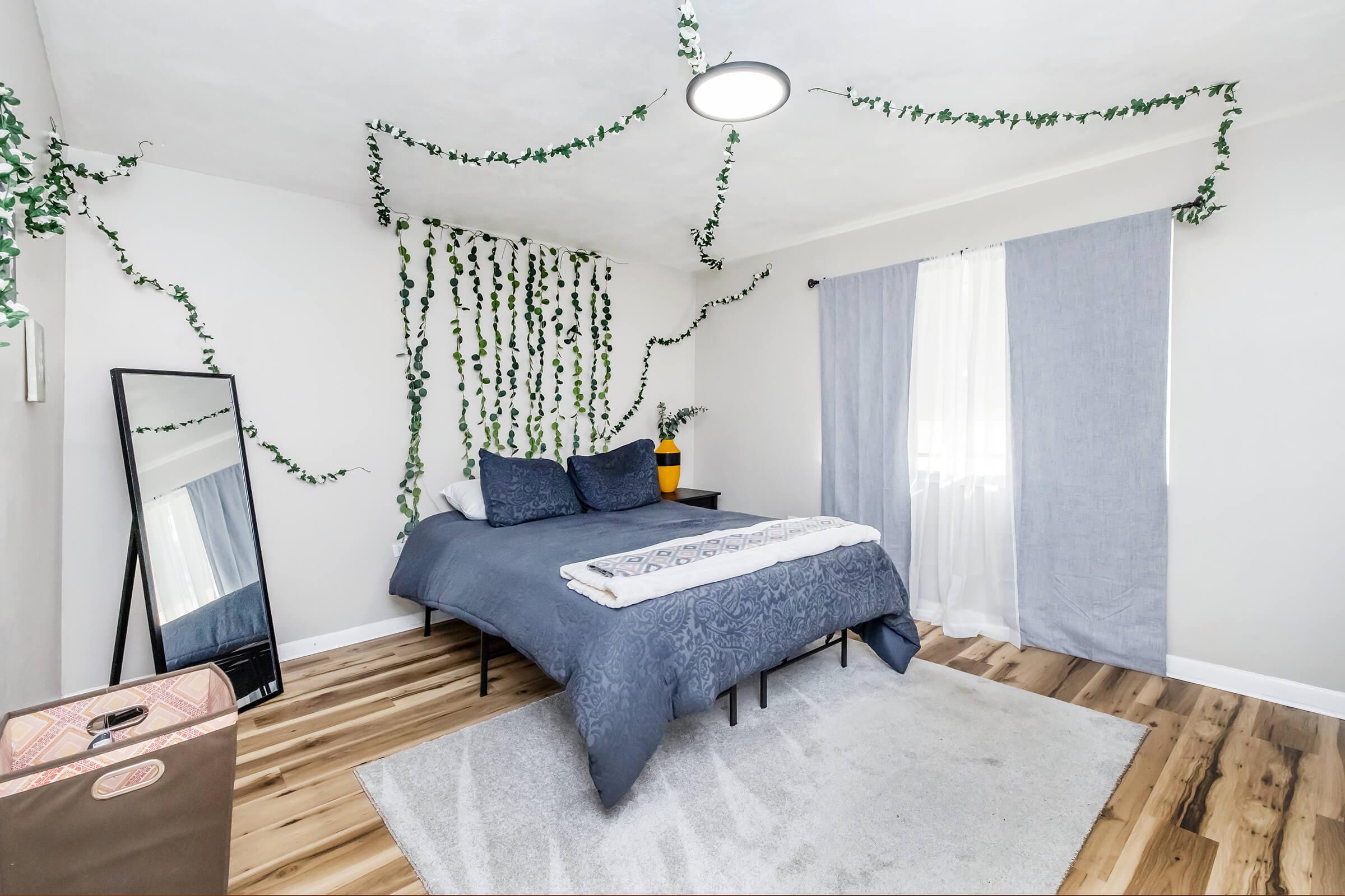 A cozy bedroom featuring a comfortable bed with blue bedding and decorative pillows. The wall is adorned with green ivy vines, and there is a large mirror beside the bed. A window with light gray curtains lets in natural light, and a soft rug lies on the wooden floor, creating a welcoming atmosphere.