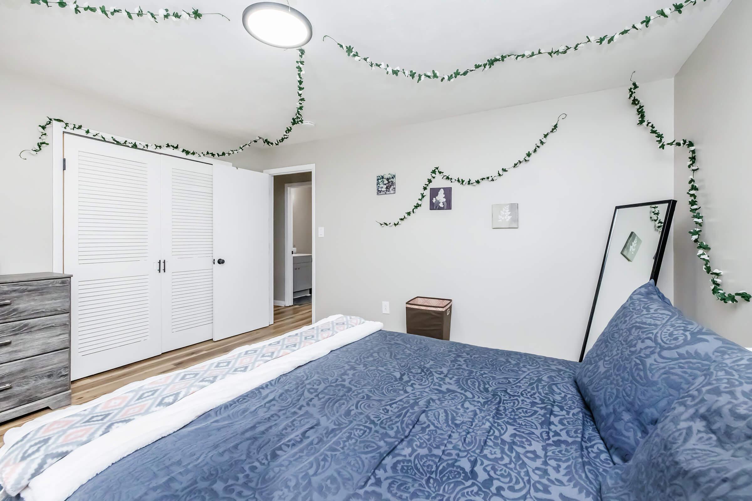 A cozy bedroom featuring a neatly made bed with a dark blue patterned comforter. Green ivy garlands are draped along the walls and ceiling. A full-length mirror and a dresser are visible. The room has light-colored walls and wood-look flooring, creating a warm and inviting atmosphere.