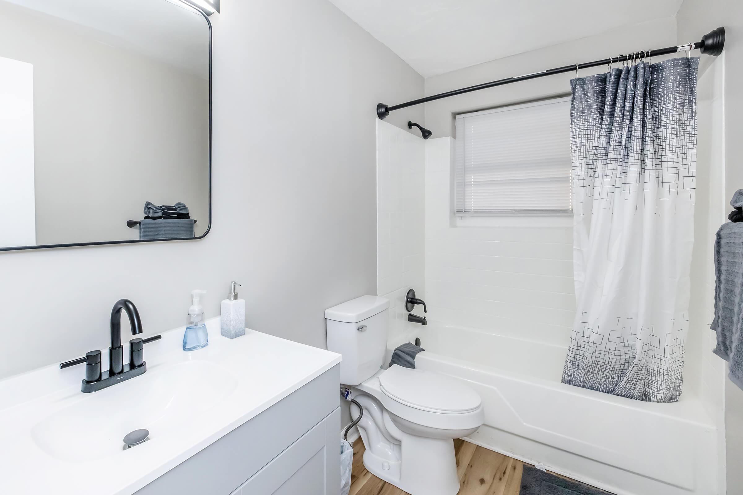 Modern bathroom with a white bathtub, a sleek black faucet, and a large mirror above a minimalist vanity. The walls are painted a soft gray, and there's a patterned shower curtain. Towels and toiletries are neatly arranged, creating a clean and inviting atmosphere. 