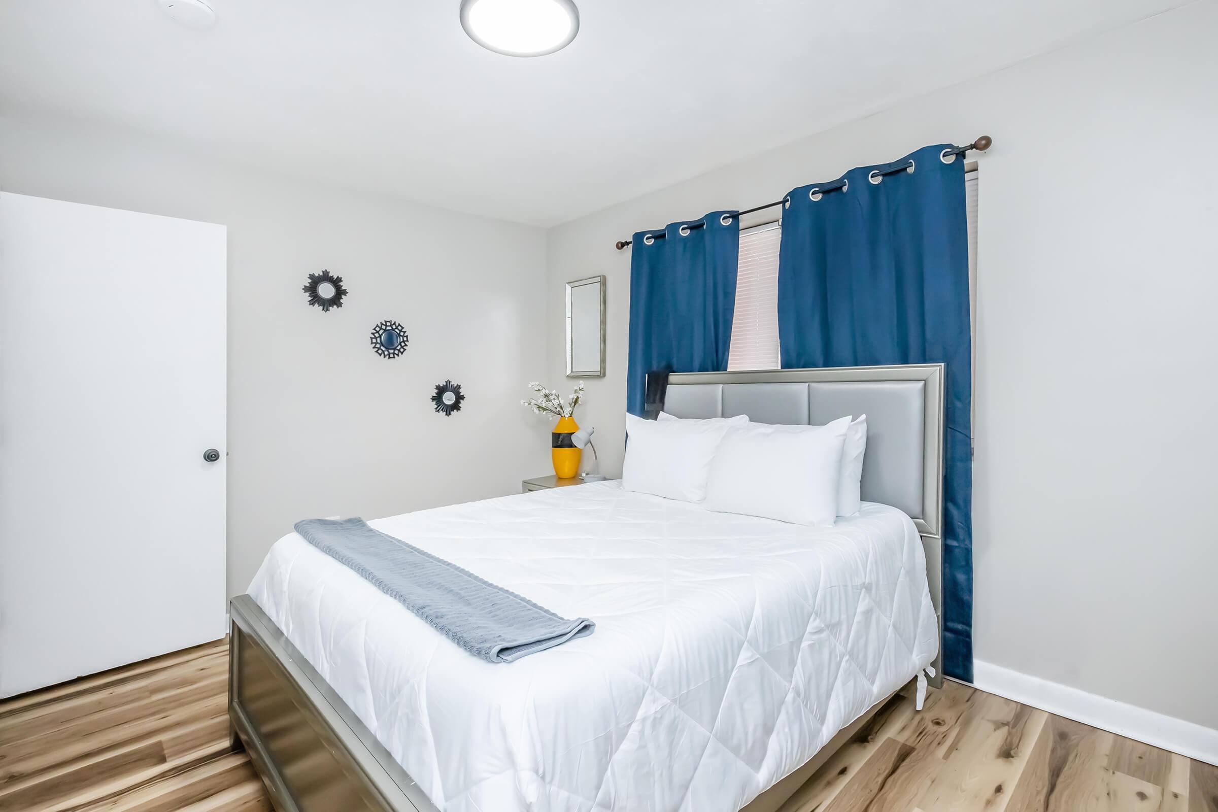 A cozy bedroom featuring a queen-sized bed with white bedding, a gray throw blanket, and two white pillows. The walls are painted light gray, adorned with wall decor. A large window is dressed with blue curtains. A small mirror and a vase with flowers are on the bedside table, and the flooring is wooden.