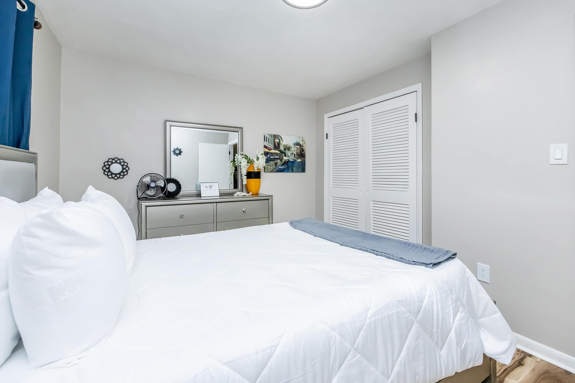A cozy bedroom featuring a neatly made bed with white linens and a gray throw blanket. There is a dresser with a mirror, a piece of wall art, and a closet with sliding doors. Soft lighting enhances the inviting atmosphere, while a fan adds a practical touch. Blue curtains frame the window, completing the decor.