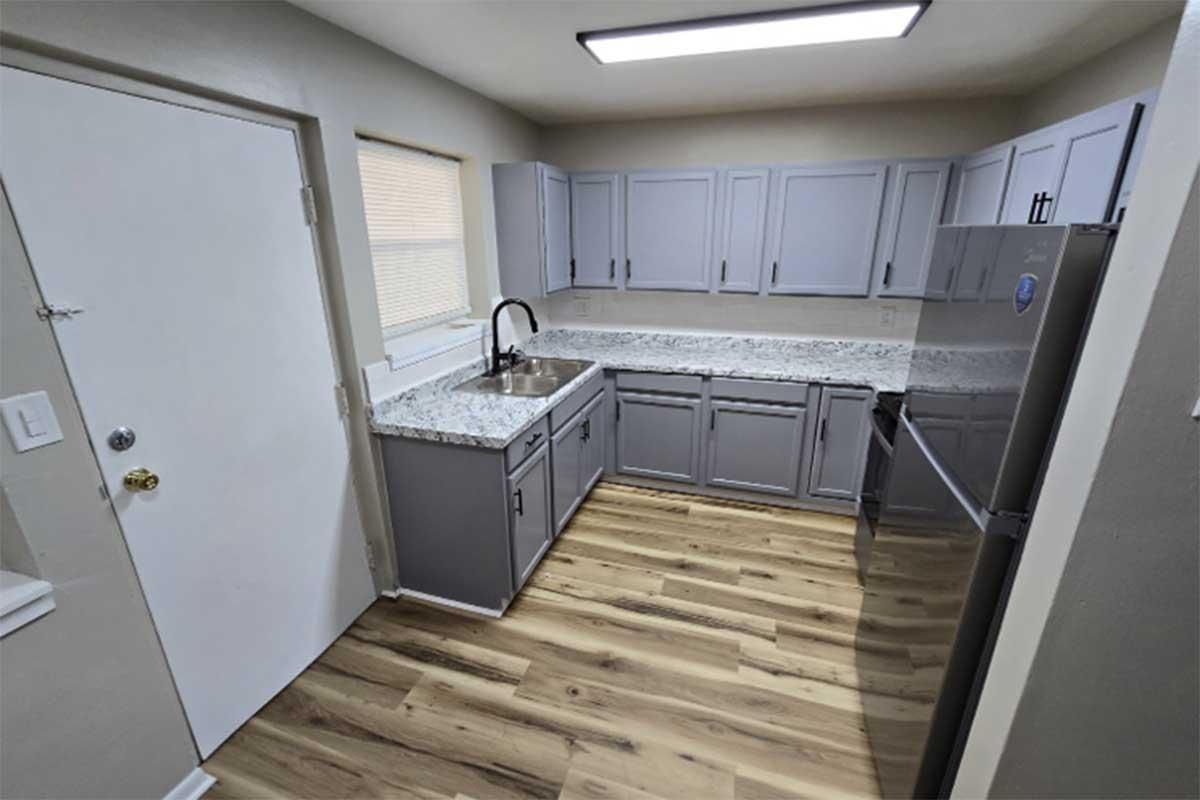 A modern kitchen featuring gray cabinetry, a granite countertop, and a stainless steel refrigerator. The room is well-lit with a ceiling light and has a door leading outside. The floor is made of light wooden planks, and there's a window above the sink allowing natural light to enter.