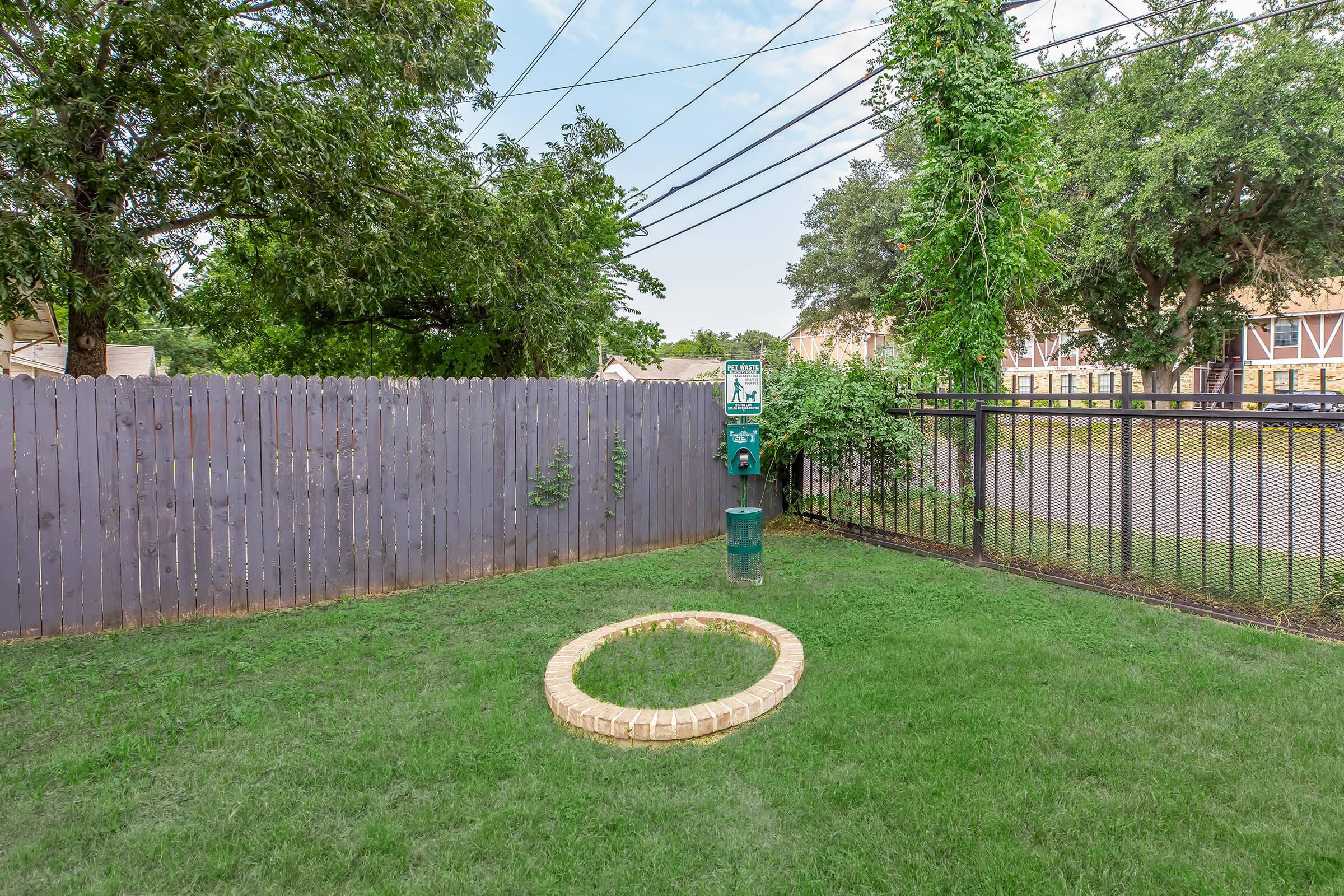 A fenced outdoor area featuring a circular brick-bordered space on green grass. In the background, there is a gray wooden fence, trees, and utility wires overhead. The setting appears to be residential, with nearby buildings visible beyond the fence.