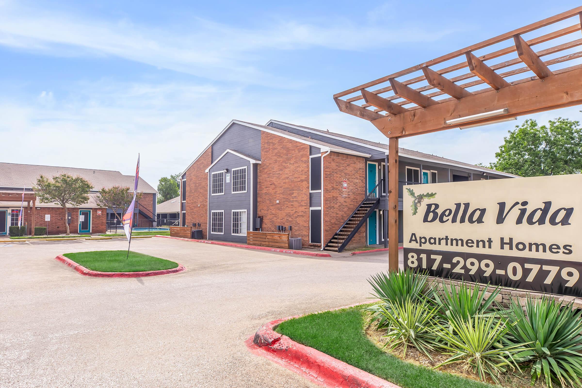 A view of the Bella Vida Apartment Homes complex, featuring brick buildings, a landscaped entrance with a sign displaying the name and contact number, and a well-maintained parking area. Nearby, there are decorative trees and plants, set against a clear sky.