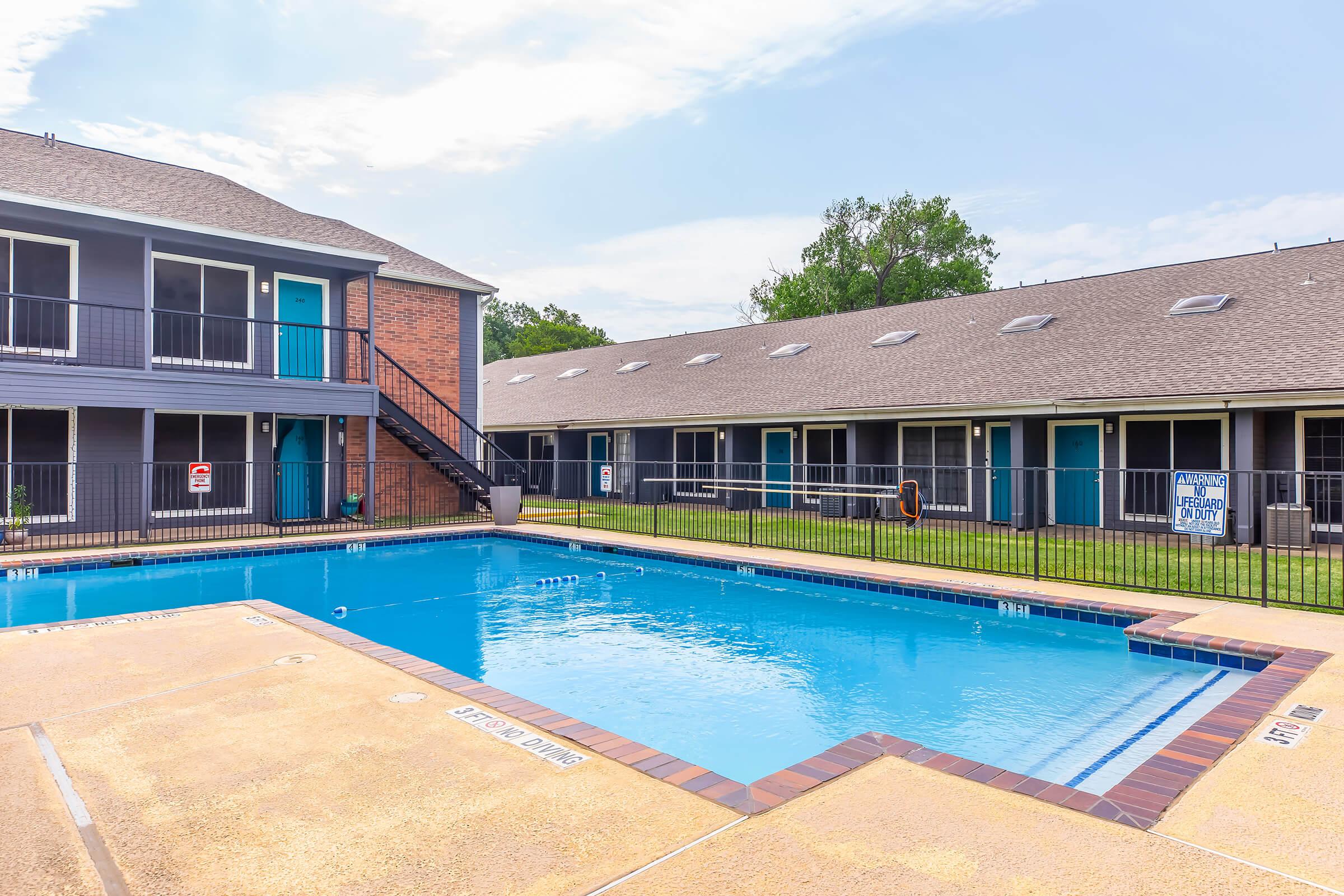 A clean swimming pool surrounded by a fenced area, with two multi-story brick and blue buildings in the background. The pool area features a concrete deck and lounge area, with clear blue water and a few poolside amenities. Bright sky and greenery visible in the background.