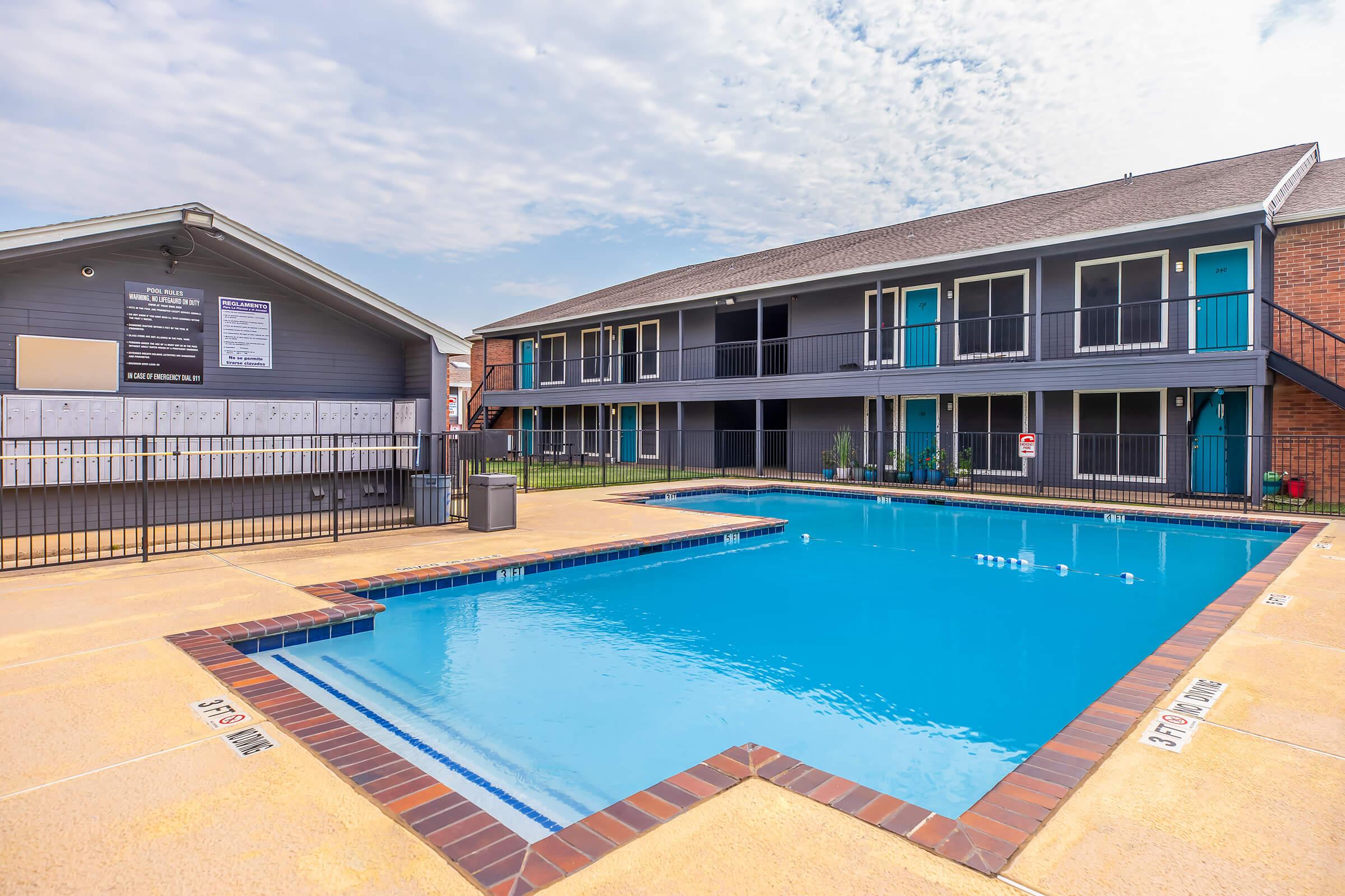 A residential pool area featuring a clear blue swimming pool surrounded by a tan deck. In the background, there are two stories of apartment buildings, with a laundry room to the left. The sky is partly cloudy, creating a calm atmosphere.