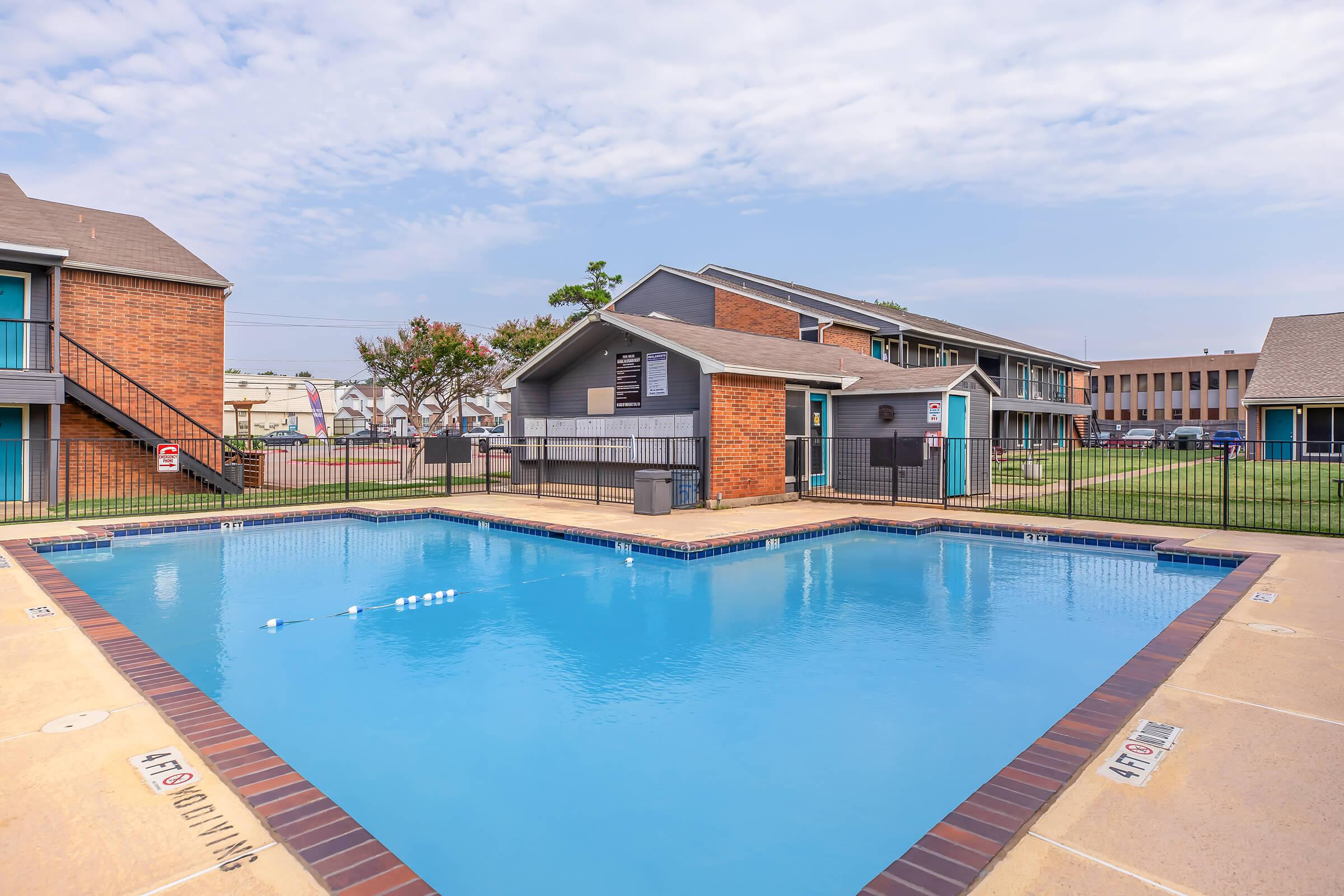 A clear blue swimming pool surrounded by a paved area, with two-story brick buildings in the background. The pool area is fenced and includes signs, with a grassy area nearby. The sky is partly cloudy.
