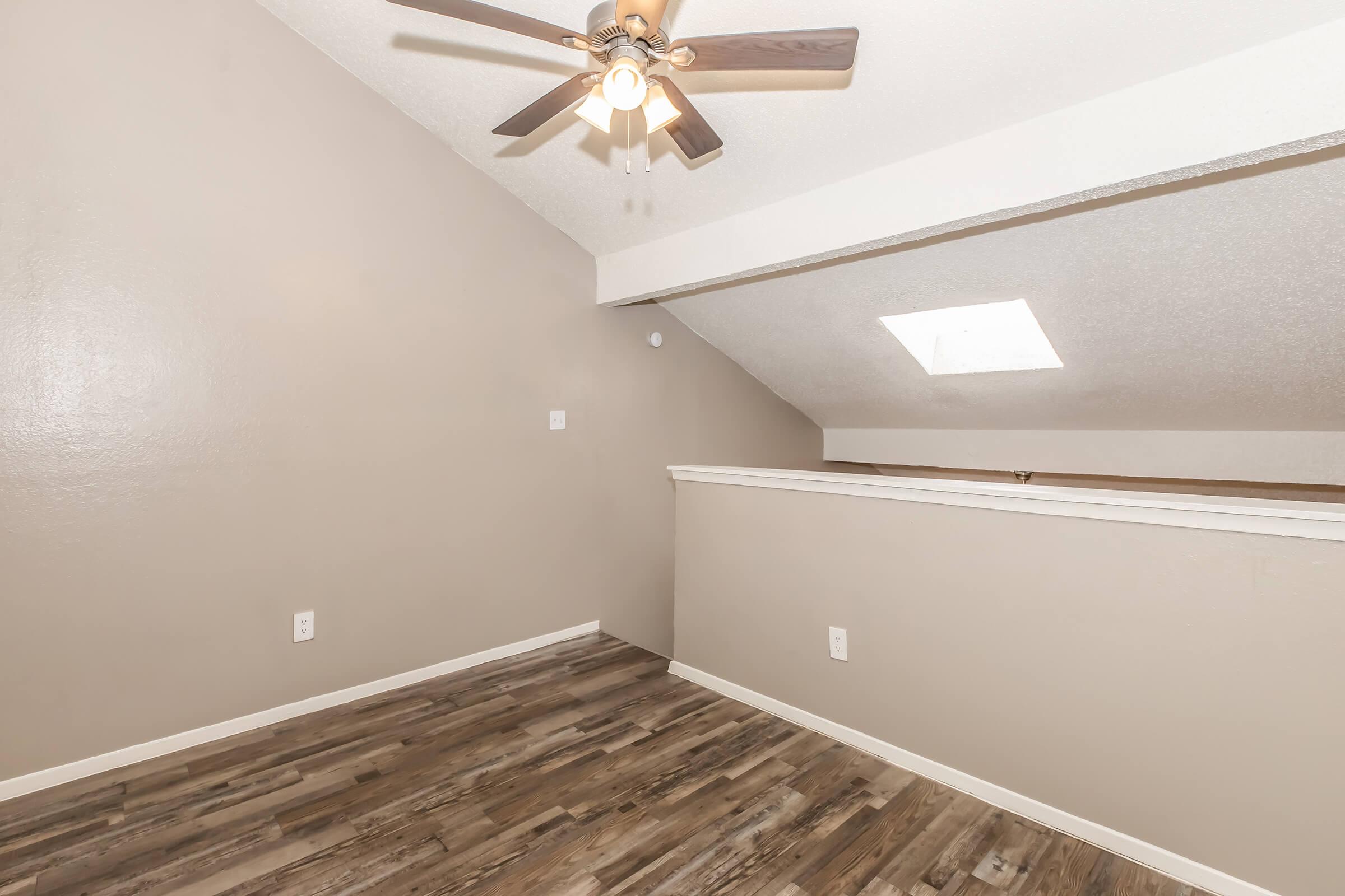Interior view of a room featuring a ceiling fan, sloped ceiling, and a skylight. The walls are painted a neutral color, and the floor is covered with wooden planks. A railing is visible in the foreground, suggesting an open design or loft space.