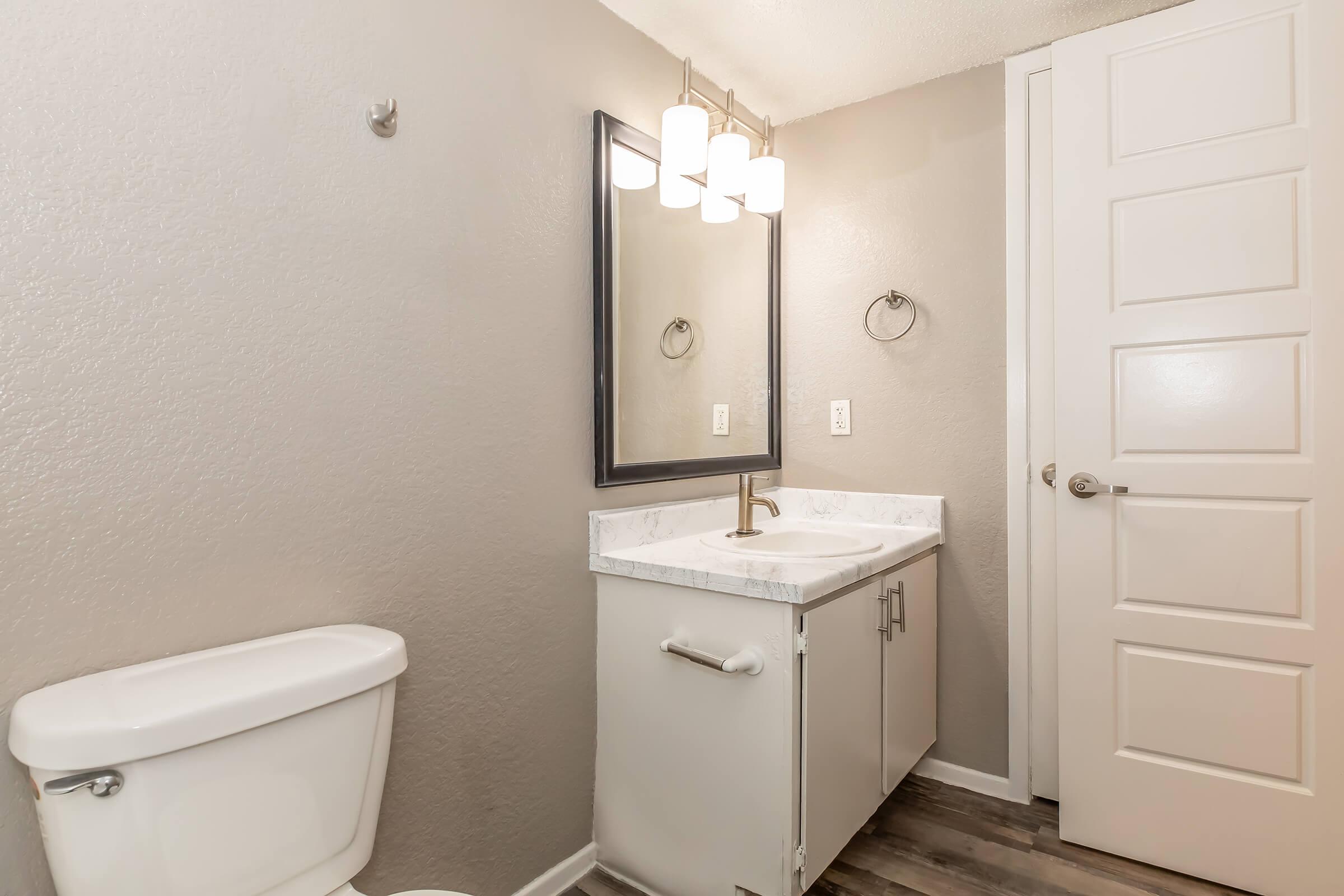 A modern bathroom featuring a white toilet, a sleek vanity with a marble countertop, and a rectangular mirror above the sink. The walls are painted a light gray, and there is a modern light fixture with three bulbs. A door is visible on the right side. The flooring is a wood-like laminate.