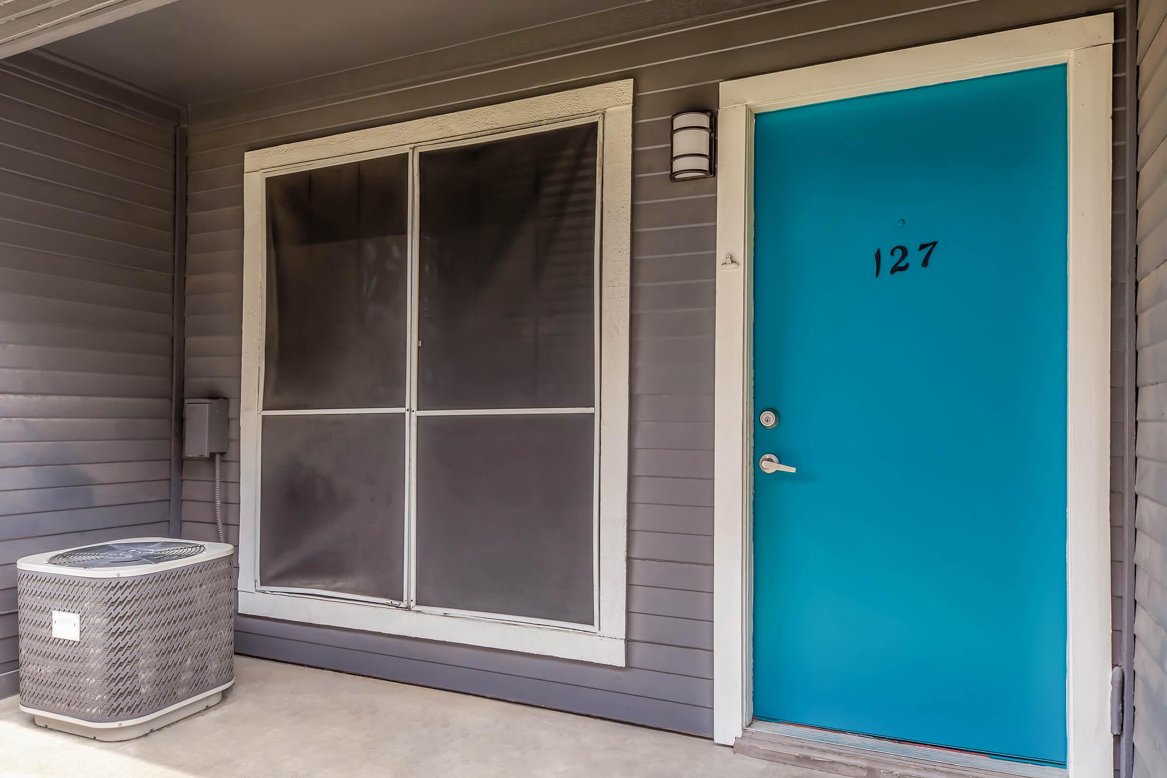 A front entrance featuring a bright turquoise door with the number 127 displayed. Next to the door is a large window covered with a black screen. An air conditioning unit is installed nearby, against a gray wall. The porch is concrete and has a modern, clean appearance.