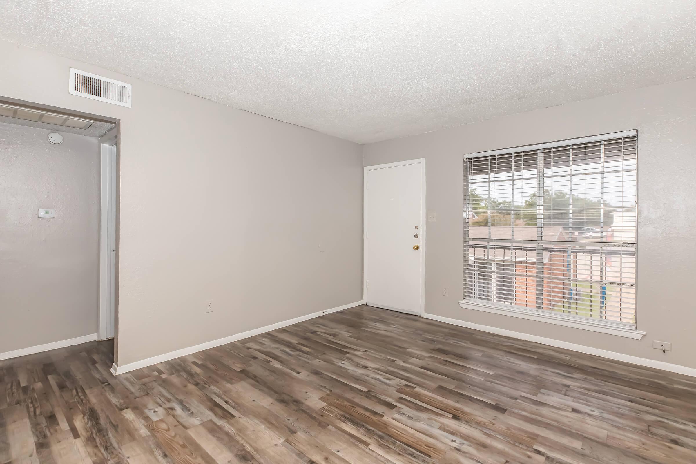 A spacious, blank room with light gray walls and a shiny wood-like floor. The room features a large window with white blinds and a front door. There is a small area with a closet or storage space visible on the left side. The overall feeling is clean and minimalistic, suitable for various uses.