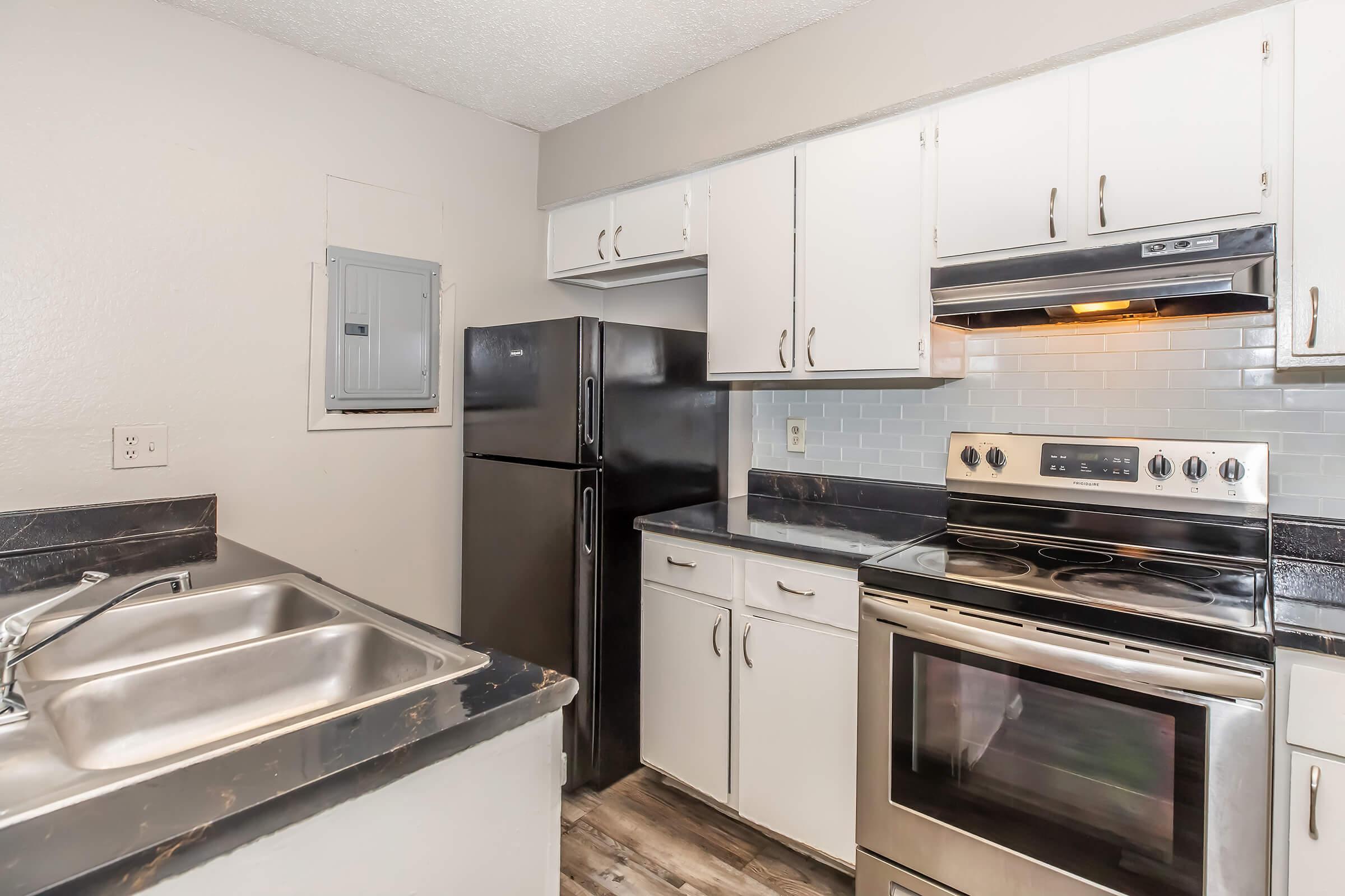 A modern kitchen featuring white cabinets, a black refrigerator, a stainless steel stove and oven, a silver sink, and a dark countertop. The walls are light-colored, and there's an electrical panel visible in the corner. Overall, the kitchen has a clean and functional design.