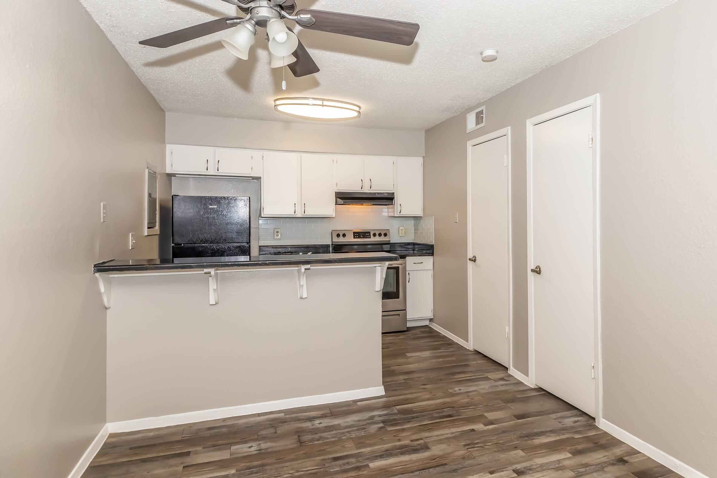Modern kitchen with a ceiling fan, stainless steel appliances, and granite countertops. Features white cabinetry and a bar area with stools. Light wood-look flooring and neutral-colored walls create a bright, inviting space. Two doors lead to different rooms, enhancing functionality.