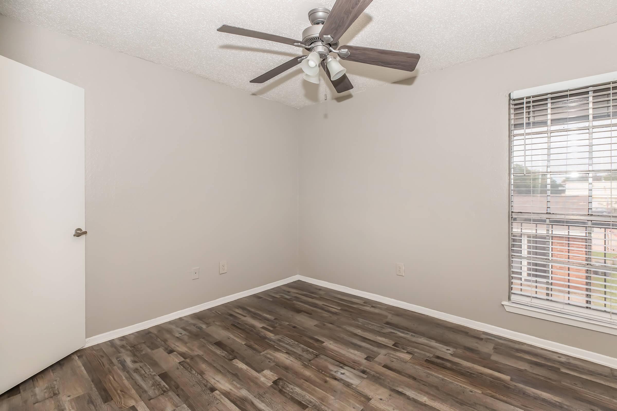 A small, empty bedroom featuring light gray walls, a ceiling fan with wooden blades, and a window with blinds. The floor is made of wood-like planks, and the door is partially open, revealing an entryway. The overall space is bright and minimalistic, suitable for various interior designs.