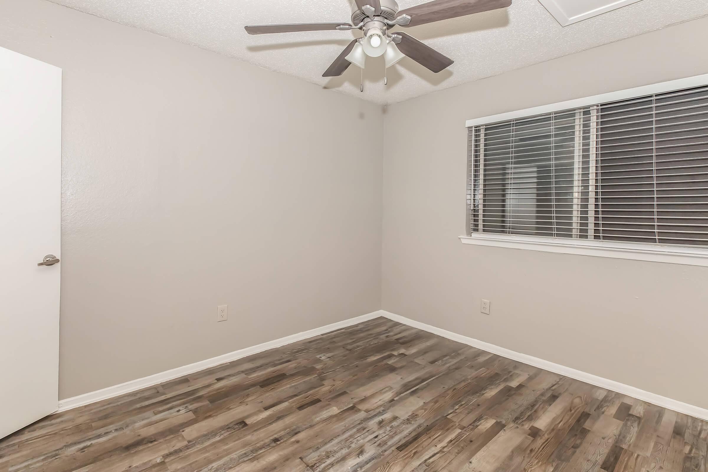 A vacant room with light gray walls, featuring a ceiling fan with lights. The floor has a wood-like finish. There's a window with white blinds on one wall and a closed door on the left side of the image. The overall atmosphere is simple and uncluttered, suitable for various uses.