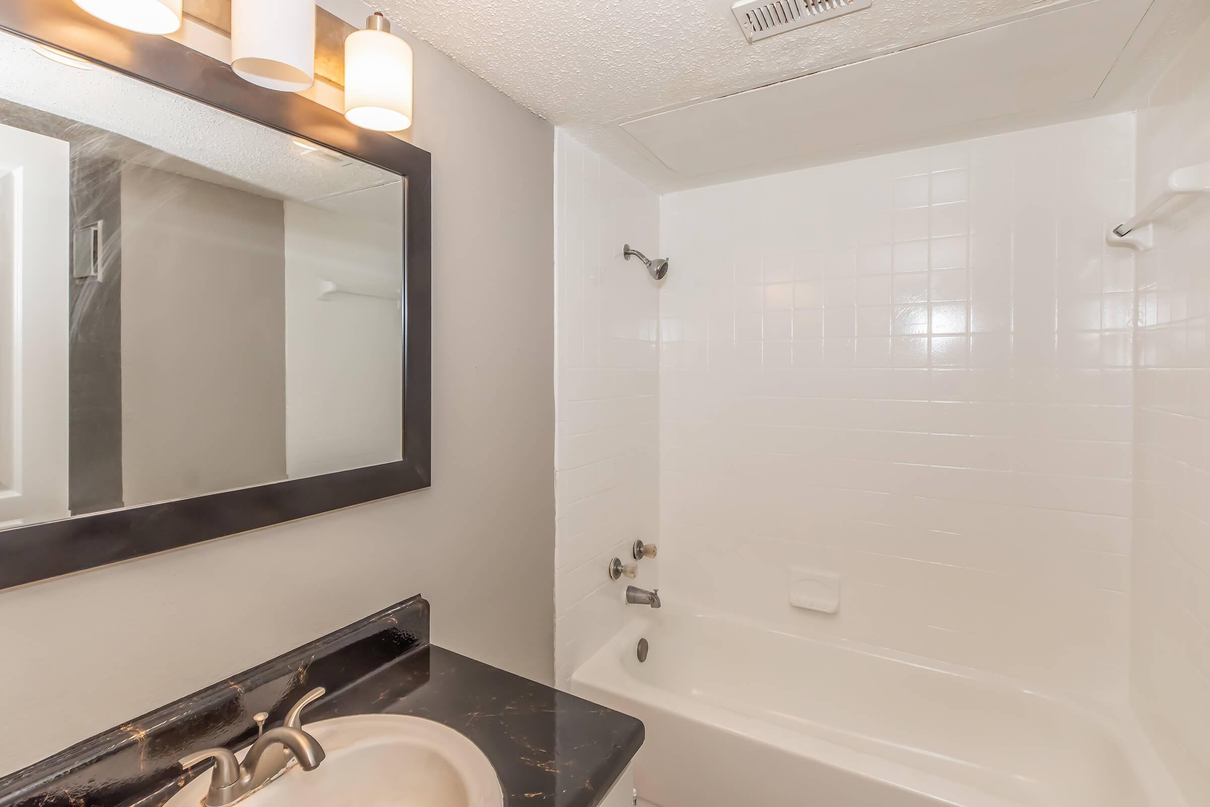 A clean, modern bathroom featuring a white tiled shower and bathtub. The space includes a dark countertop with a sink and a large mirror above it. Three light fixtures are mounted above the mirror, providing bright illumination. The walls are painted in a light gray color, giving a fresh and airy look.