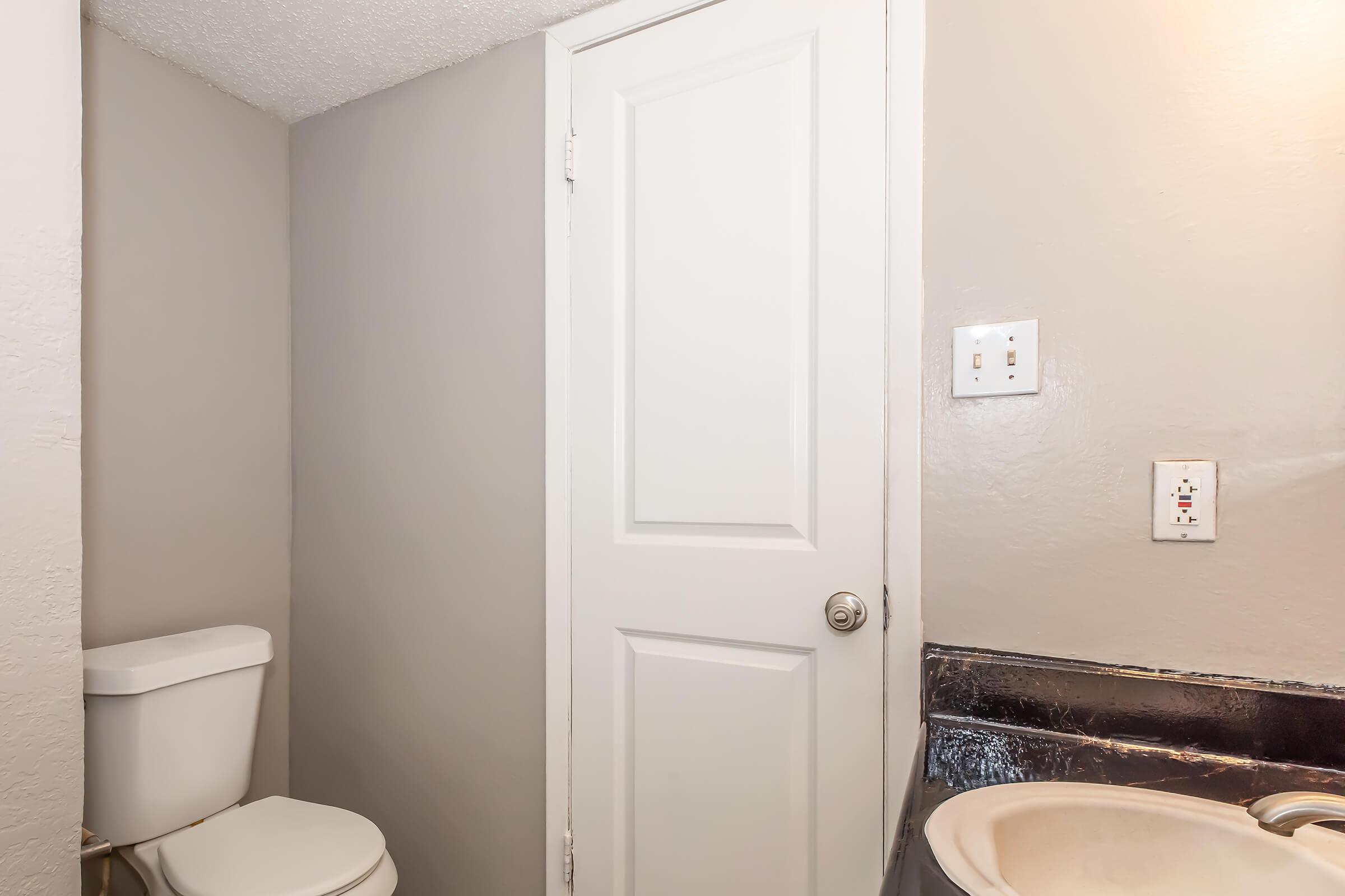Interior view of a small bathroom featuring a white toilet, a closed door, and a sink. The walls are painted light gray, and there is a countertop visible near the sink. The overall space appears compact and functional.