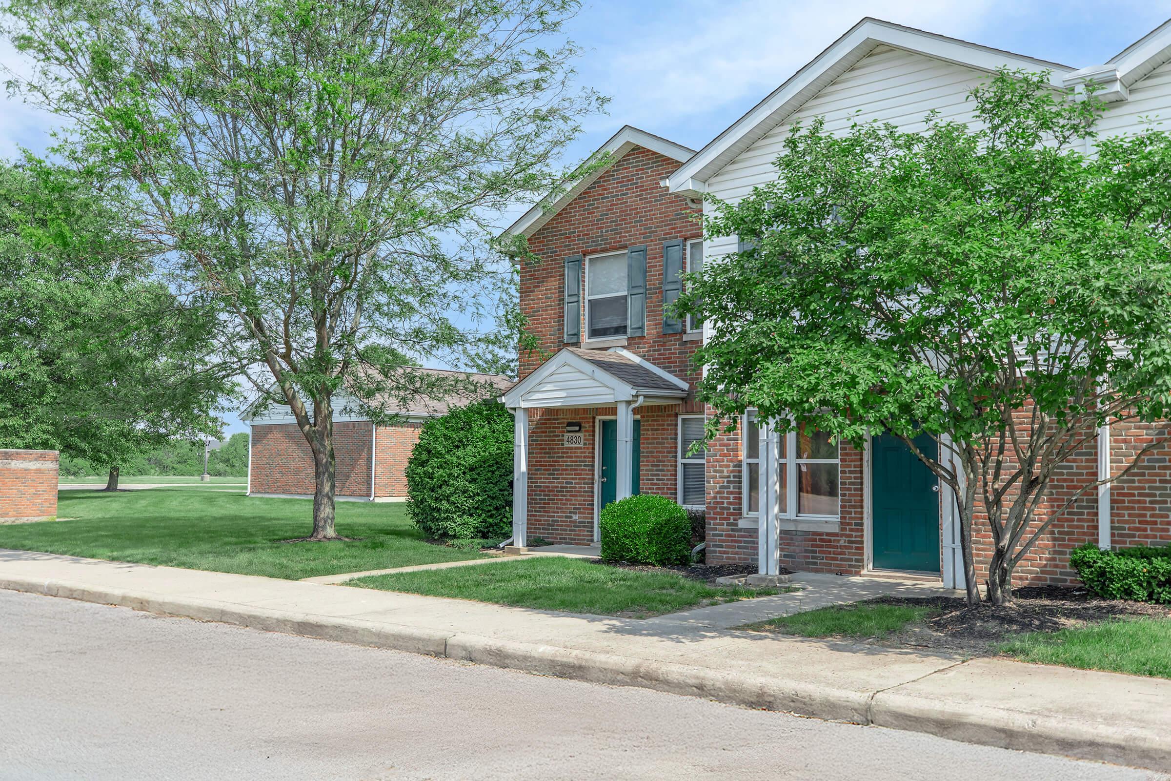 A residential building with brick exterior and green doors, flanked by shrubs and trees, situated along a paved street. Well-manicured lawns are visible in the background, under a partly cloudy sky.