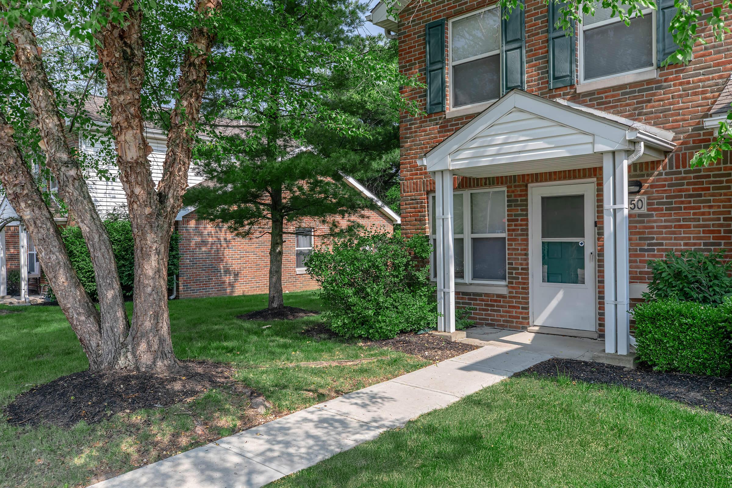 A well-maintained brick townhouse with a white front porch and green shutters. A concrete walkway leads to the entrance, bordered by neatly trimmed grass and shrubs. To the left, there's a tree providing shade. The setting is sunny and inviting, showcasing a peaceful residential atmosphere.
