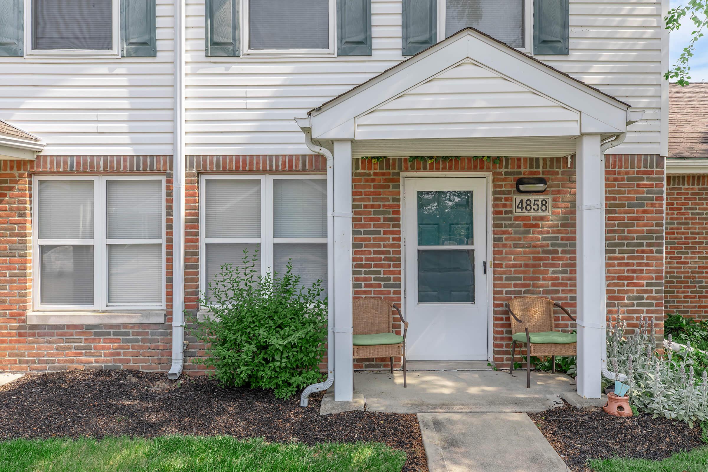 A welcoming entryway to a residential building featuring a white front door with a small overhang, flanked by two chairs on either side. The exterior has red brick and wood siding, with green bushes and grass in the foreground. A house number, 4858, is visible beside the door.