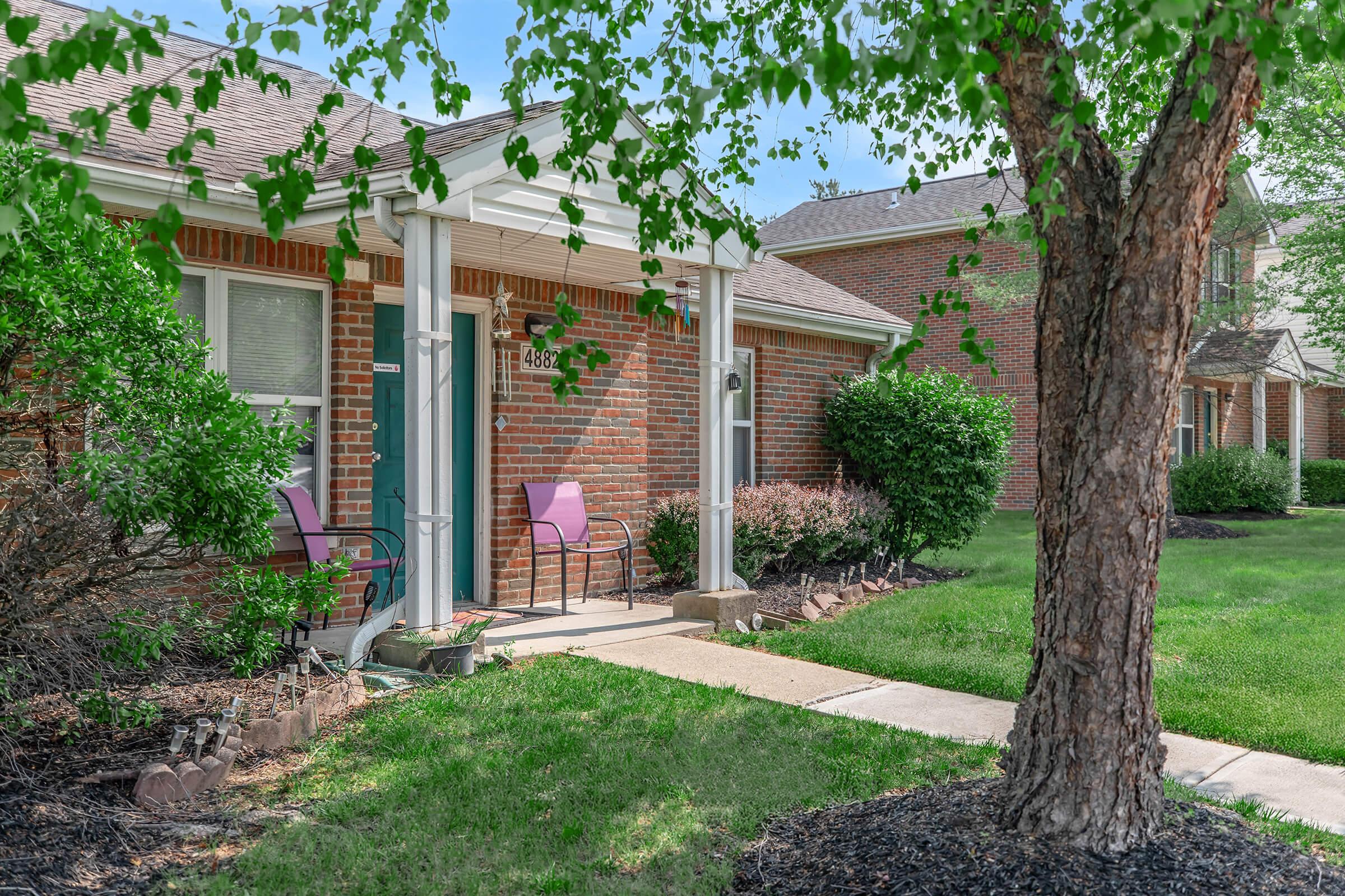 A well-maintained residential entrance featuring a brick facade, two pink chairs, and lush greenery. The sidewalk leads to a door, with bushes and trees framing the yard, creating a welcoming atmosphere in a suburban setting.