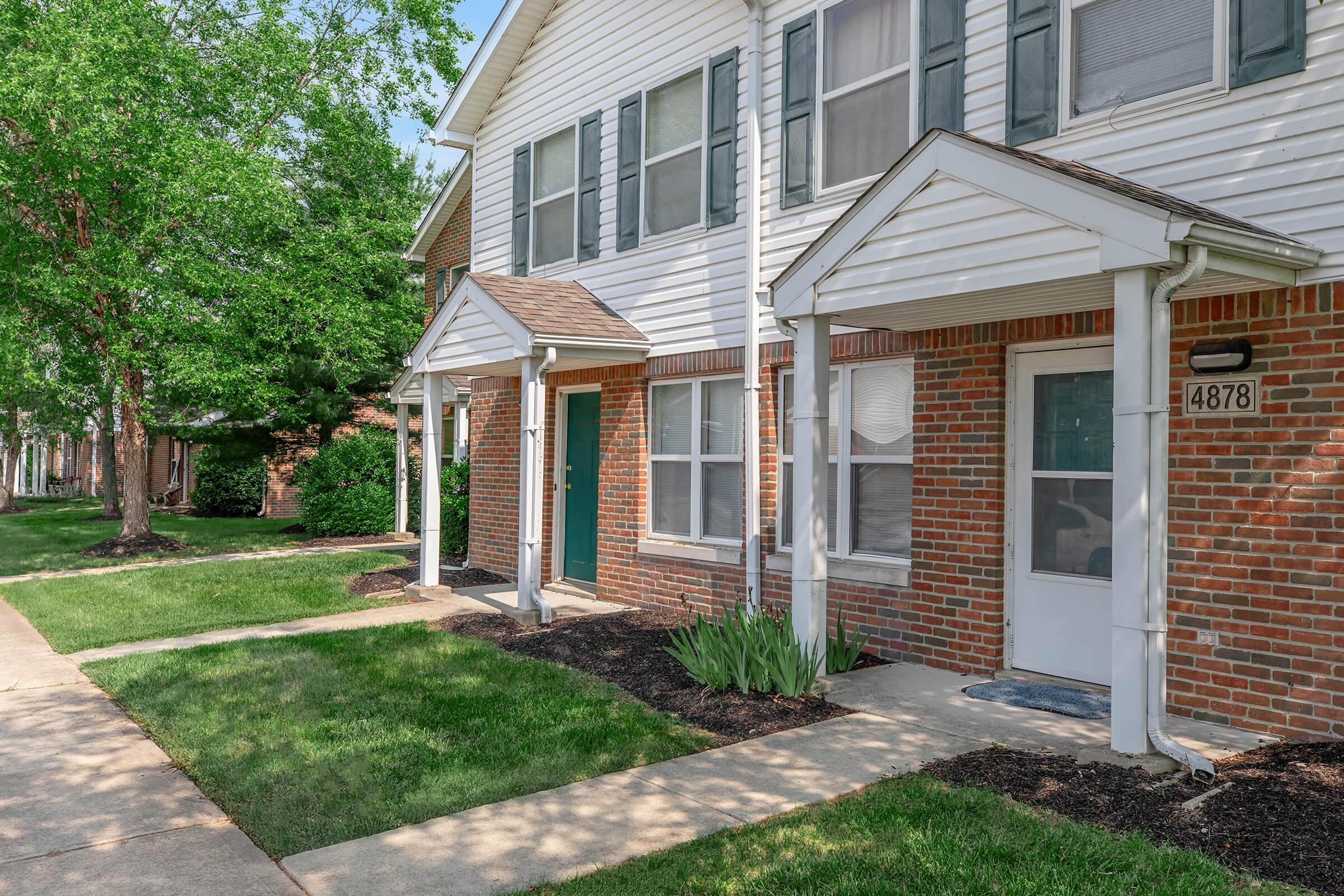 A row of townhouse entrances with brick exteriors and white trim, featuring small porches with peaked roofs. Lush green grass lines the pathways, and trees provide shade in the background. The house numbers are visible, with one door painted a contrasting color. The setting is bright and well-maintained.