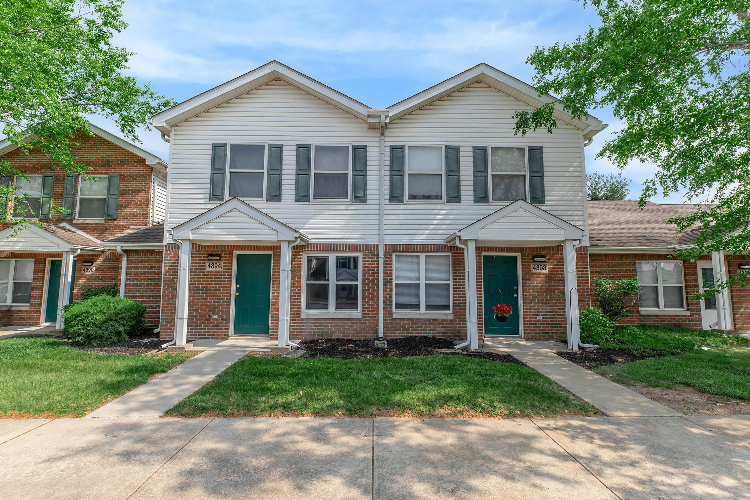 Two identical two-story townhouses with brick facades and white siding. Each unit features green doors and symmetrical windows. The area is landscaped with green grass and small bushes, and a walkway leads up to the front doors. The sky is clear and blue, indicating a sunny day.