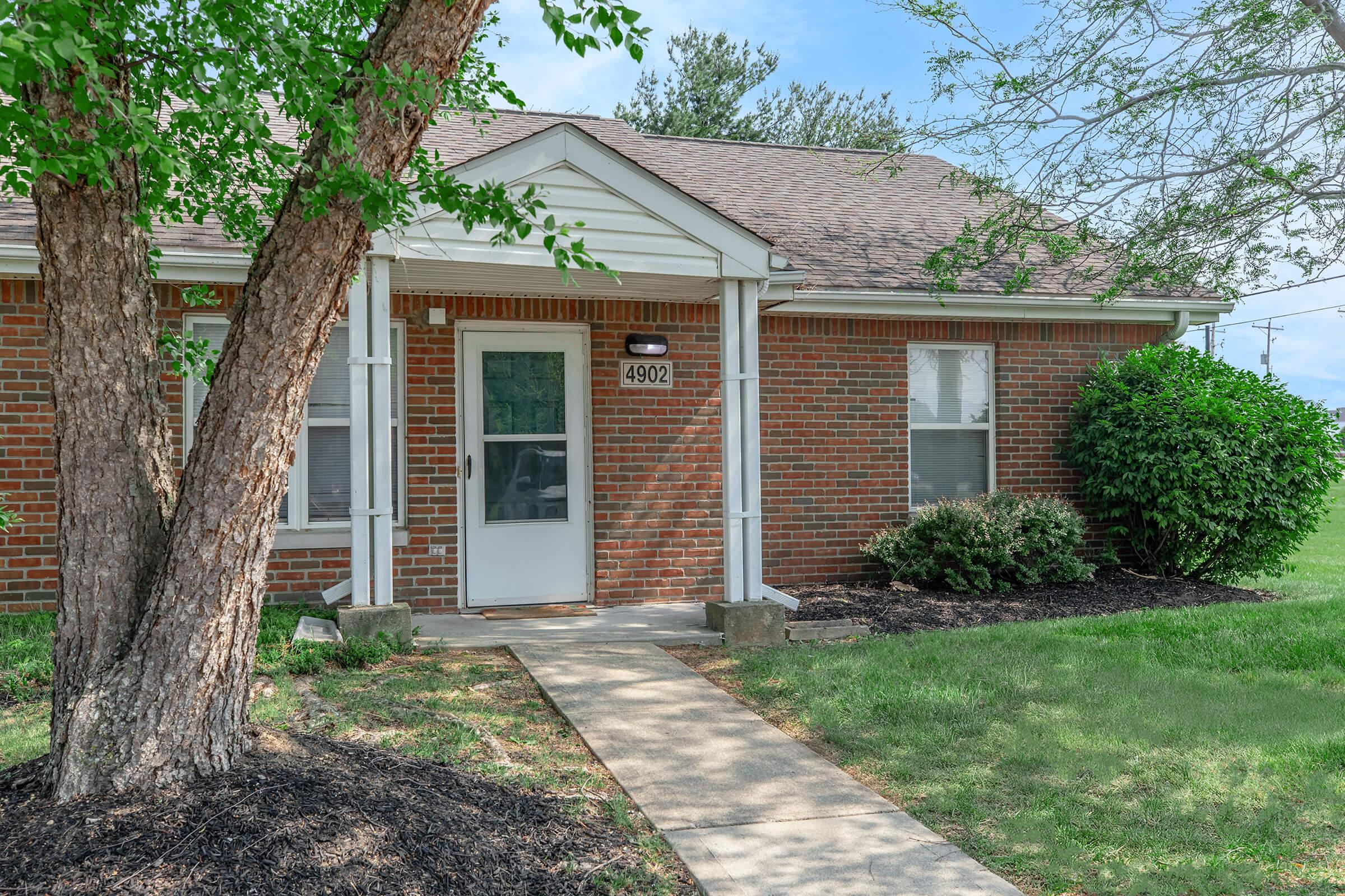 A brick house with a white front door is shown, surrounded by a lush green lawn and a small tree. The pathway leading to the door is lined with mulch, and there are a couple of windows on the house. The sky is partly cloudy, providing a bright backdrop to the scene.