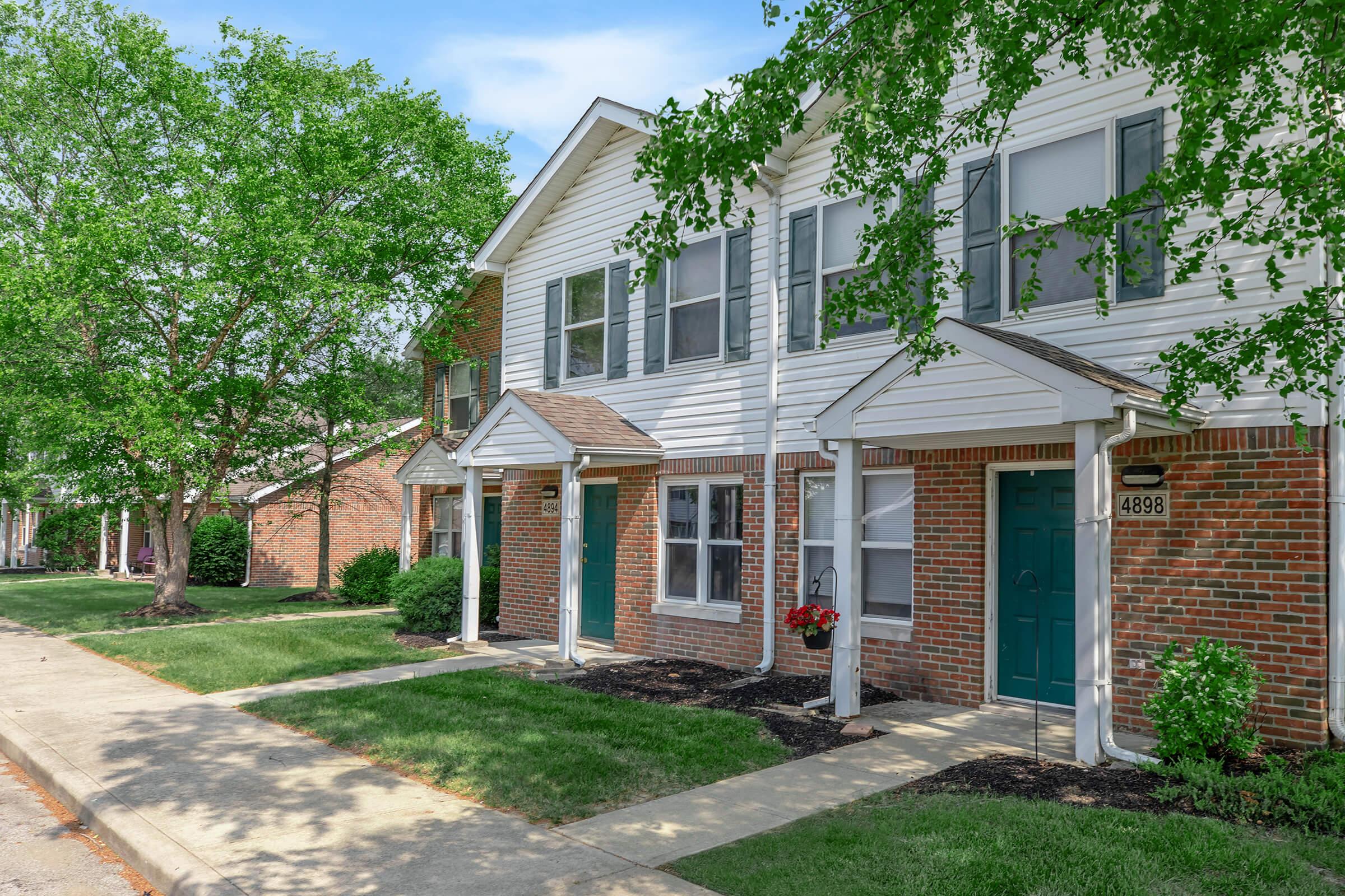 Row of townhouse-style apartments with brick exteriors and white trims. Each unit has a front door with a small covered entry and a window beside it. Lush green grass and shrubs surround the buildings, with trees providing shade. The street is paved and leads up to the homes. Bright, sunny sky in the background.