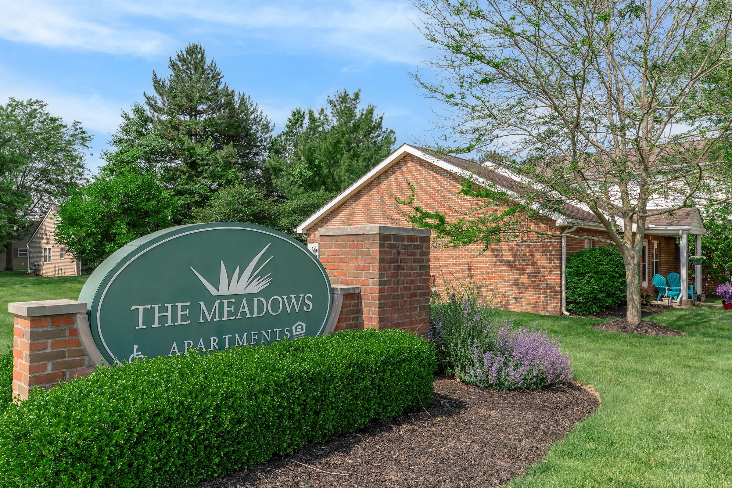 Sign for "The Meadows Apartments" surrounded by green grass, shrubs, and trees. A brick building with a welcoming atmosphere is visible in the background, along with seating areas. The scene is bright and inviting, showcasing a well-maintained residential environment.