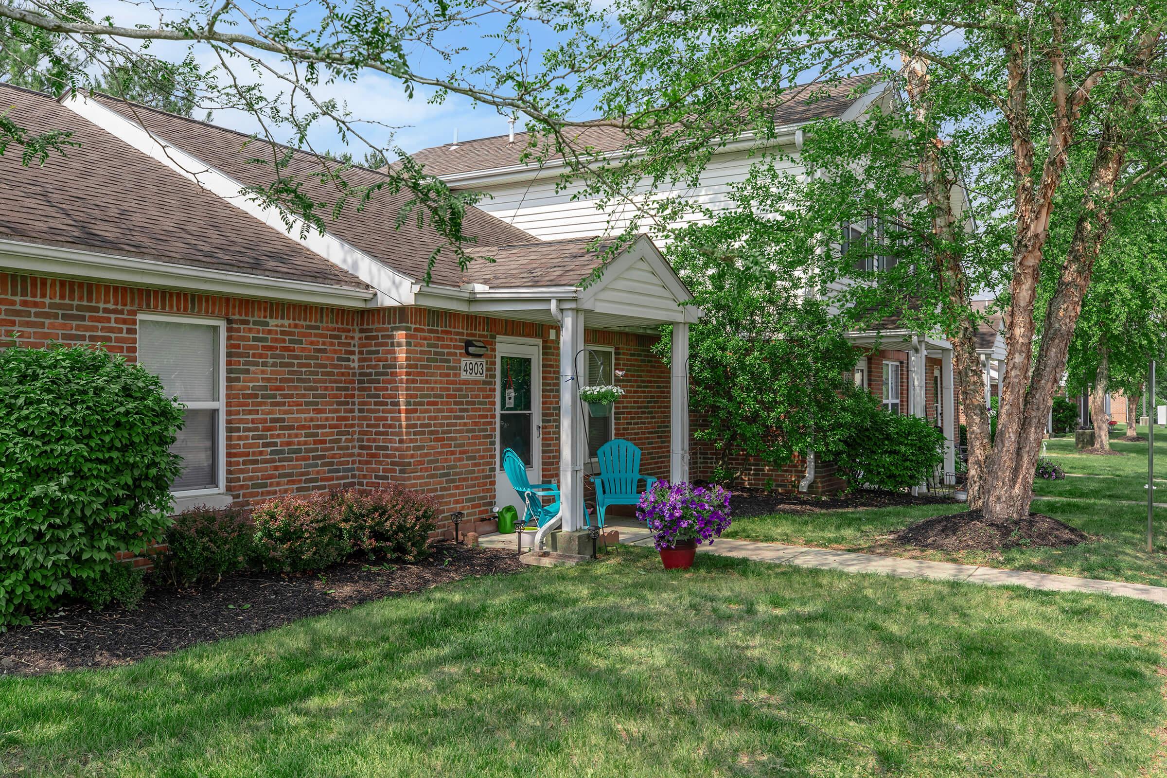 A cozy brick house with a welcoming porch featuring two blue chairs. The front yard has green grass, a flower pot with colorful flowers, and trees providing shade. Nearby, a pathway leads through the lush landscape.
