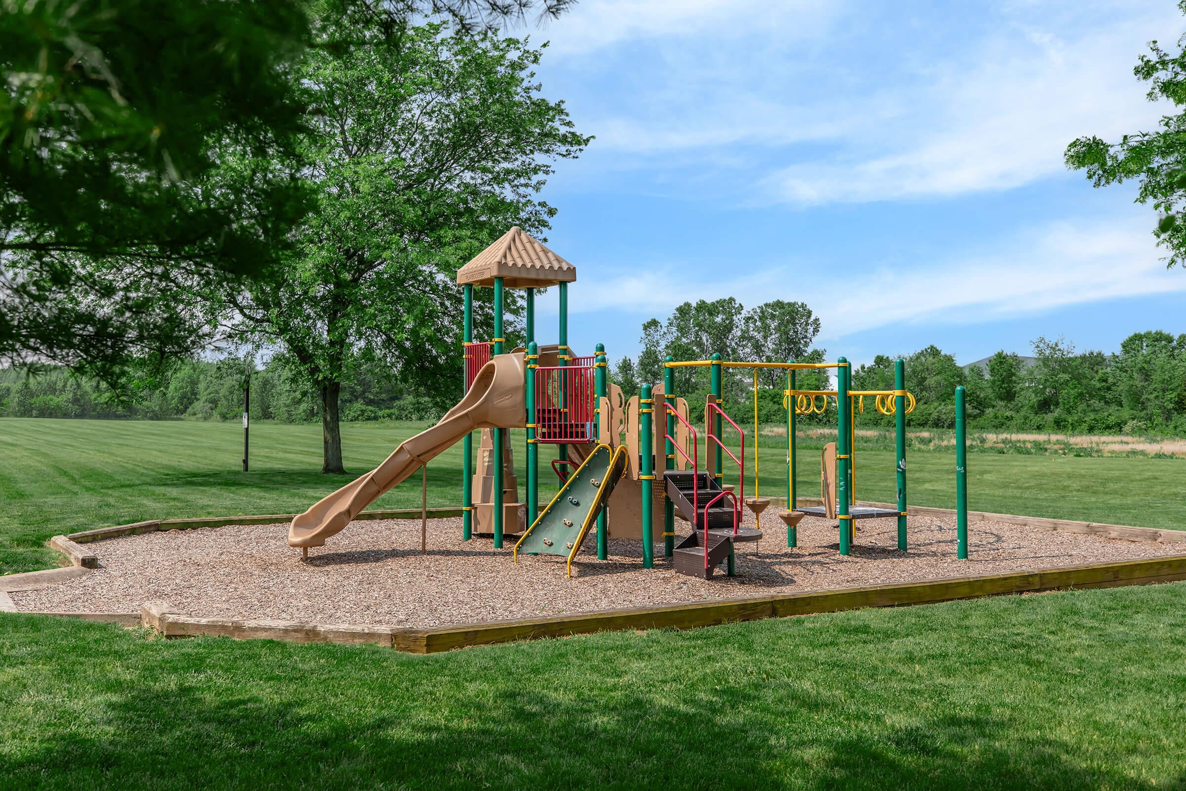 A colorful playground set in a grassy park features a slide, climbing structure, swings, and monkey bars. The area is surrounded by trees and open space, under a bright blue sky. Ideal for children to play and explore in a safe and inviting environment.