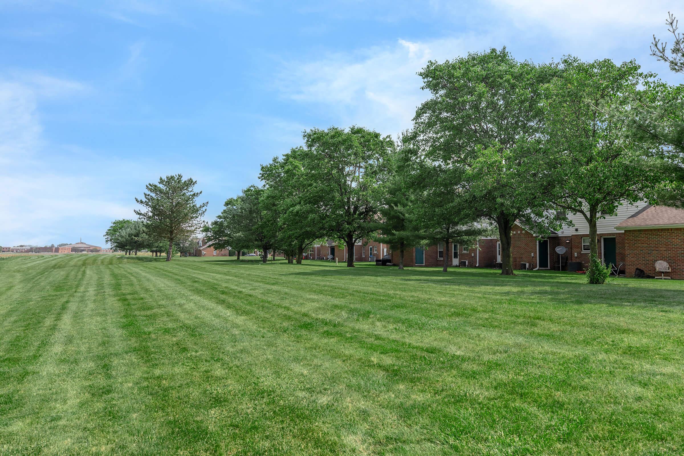 A grassy field lined with trees on one side, with brick buildings partially visible in the background under a blue sky. The scene conveys a serene and well-maintained outdoor space.