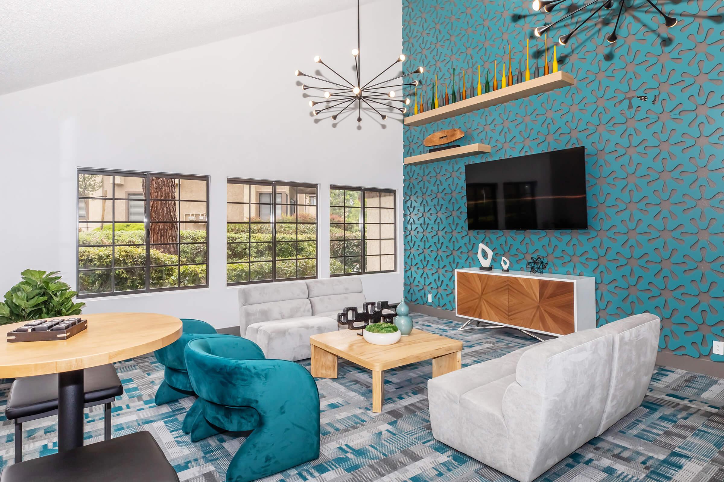 Modern living room with a unique teal accent wall featuring a geometric pattern. It includes a circular wooden table, plush gray and teal sofas, and a coffee table. Large windows allow natural light, overlooking greenery. There’s also a sleek TV unit and stylish decor items enhancing the contemporary ambiance.
