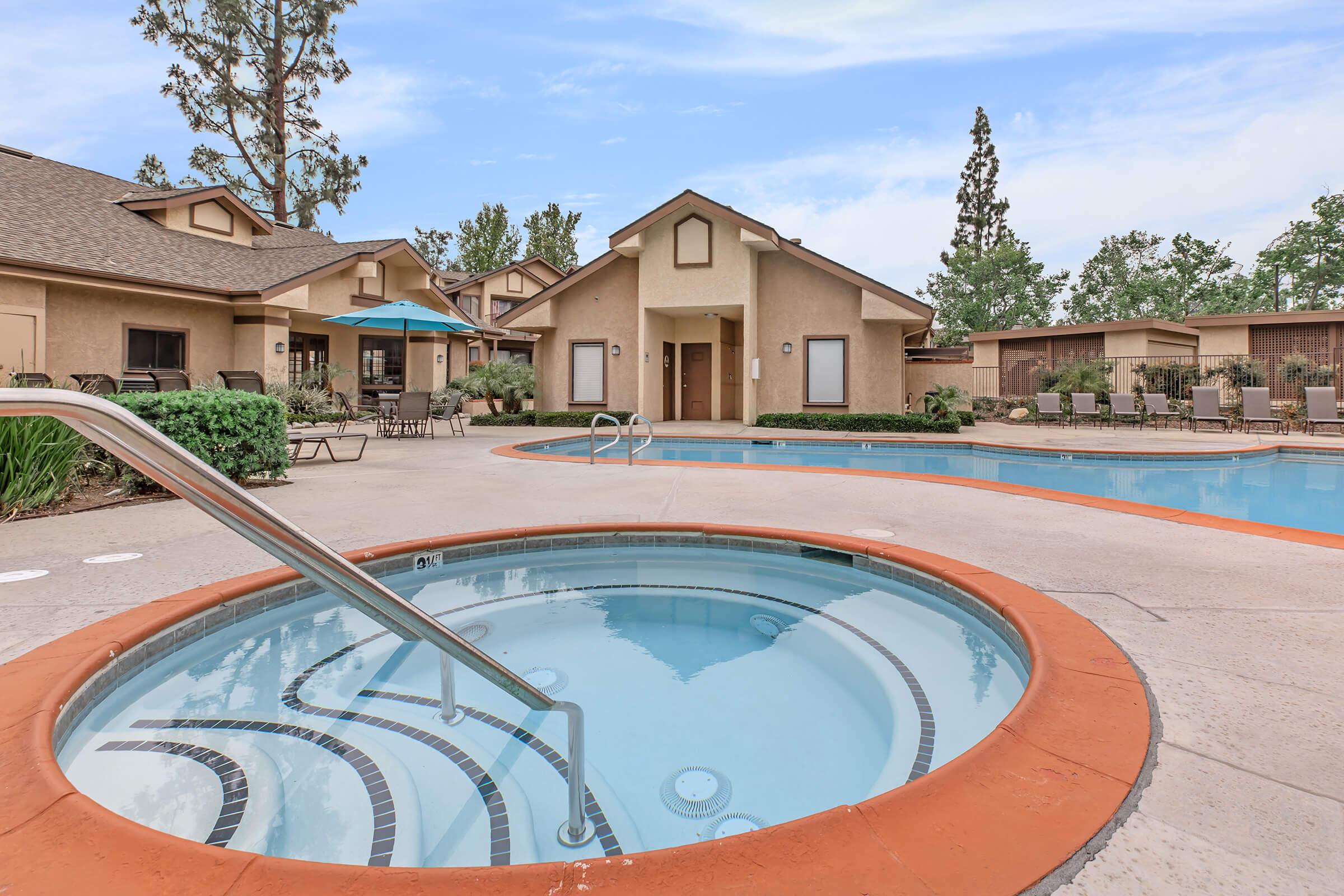 A spacious outdoor area featuring a swimming pool and a hot tub. Surrounding the pool are lounge chairs and tables, with a shaded umbrella for relaxation. Nearby, there are comfortable seating arrangements and well-maintained landscaping, creating a tranquil atmosphere for leisure and enjoyment.