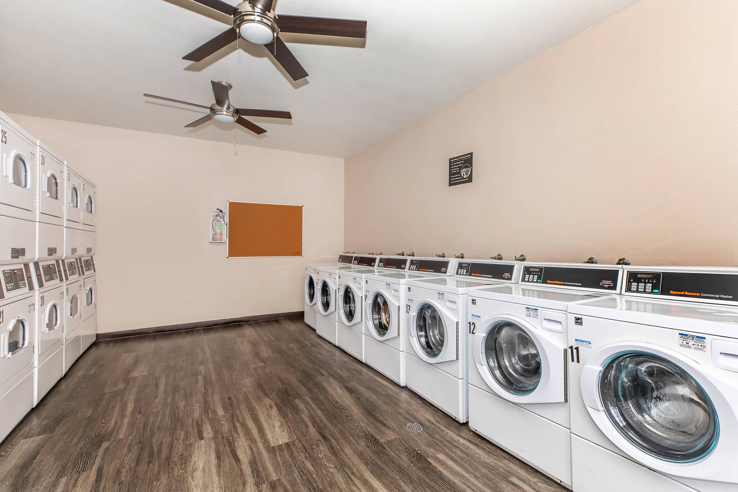 A clean and well-lit laundry room featuring several rows of white washing machines and dryers. The flooring is a dark wood laminate, and there are ceiling fans for ventilation. A bulletin board is mounted on the wall, and there is a gentle, neutral color scheme throughout the space.