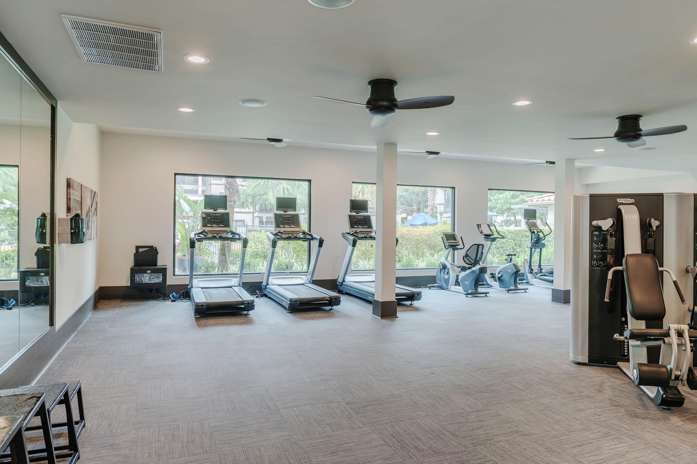 A modern gym interior featuring several treadmills, an elliptical machine, and a weight training station. Large windows allow natural light, and ceiling fans circulate air. The space has a clean, organized layout with a neutral color scheme.