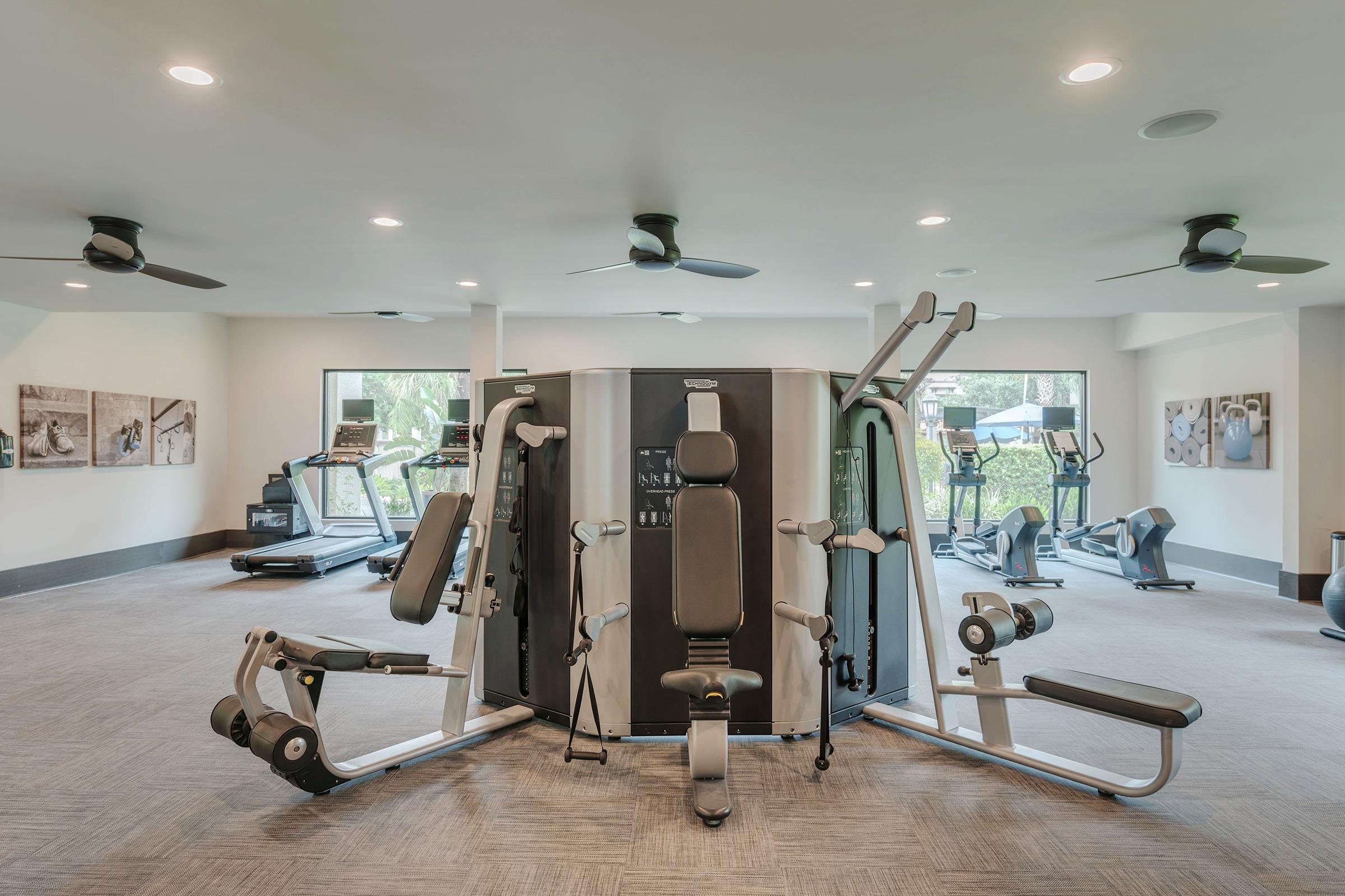 Modern gym interior featuring various fitness equipment, including a multi-station weight machine, treadmills, and exercise bikes. The space is well-lit with large windows and ceiling fans, complemented by framed artwork on the walls. The flooring is carpeted, providing a comfortable workout environment.