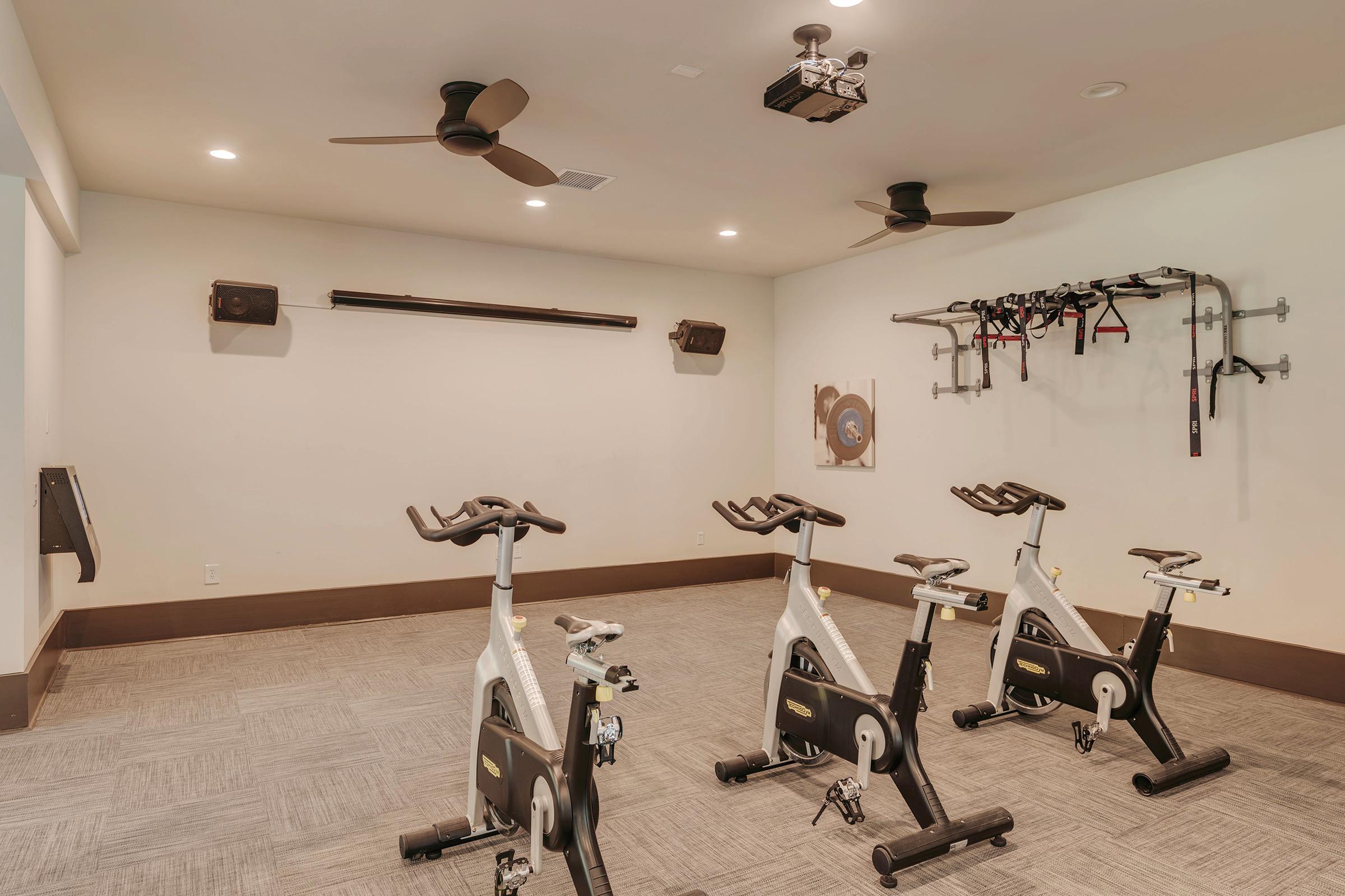 A modern exercise studio featuring four stationary bikes, equipped with ceiling fans for ventilation. The room has a light-colored wall and beige carpet, along with fitness equipment mounted on the wall and a large piece of artwork. Good lighting enhances the spacious feel of the area.