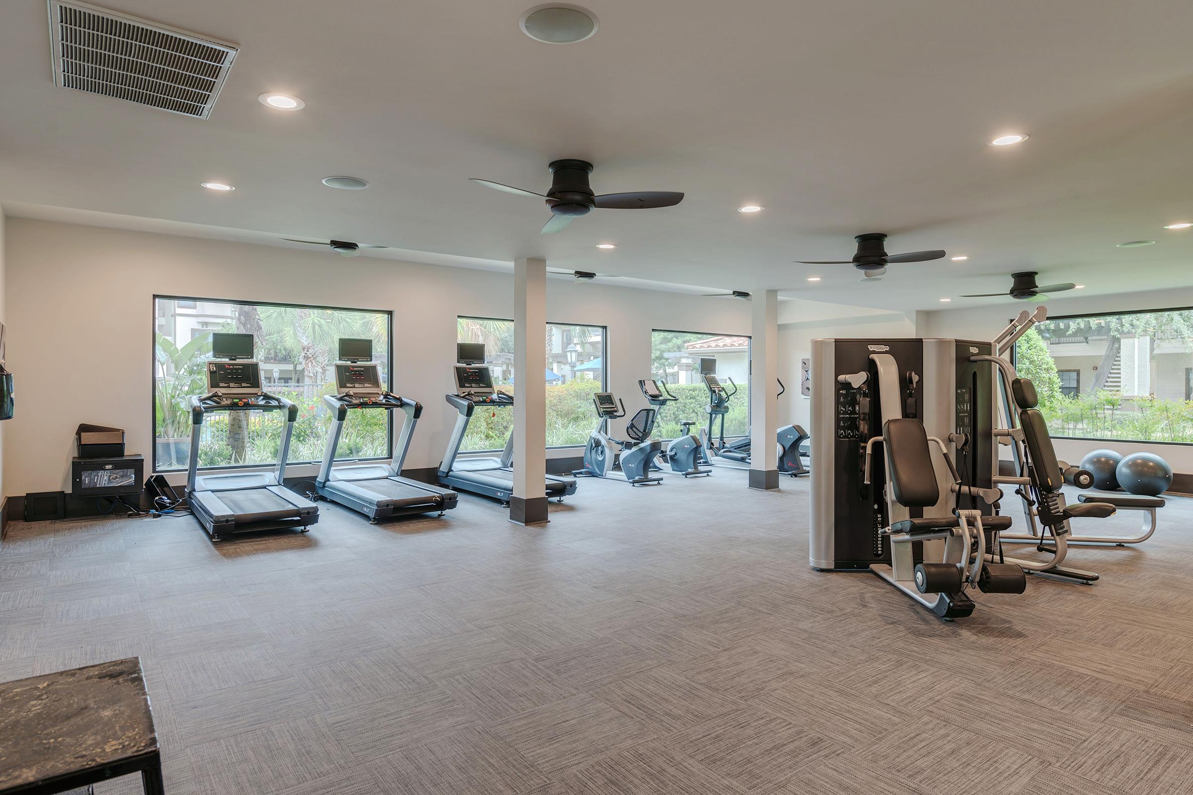 A modern gym interior featuring multiple fitness machines, including treadmills and an exercise bike. Large windows allow natural light, with views of greenery outside. The space is well-lit, clean, and organized, creating a welcoming environment for workouts.